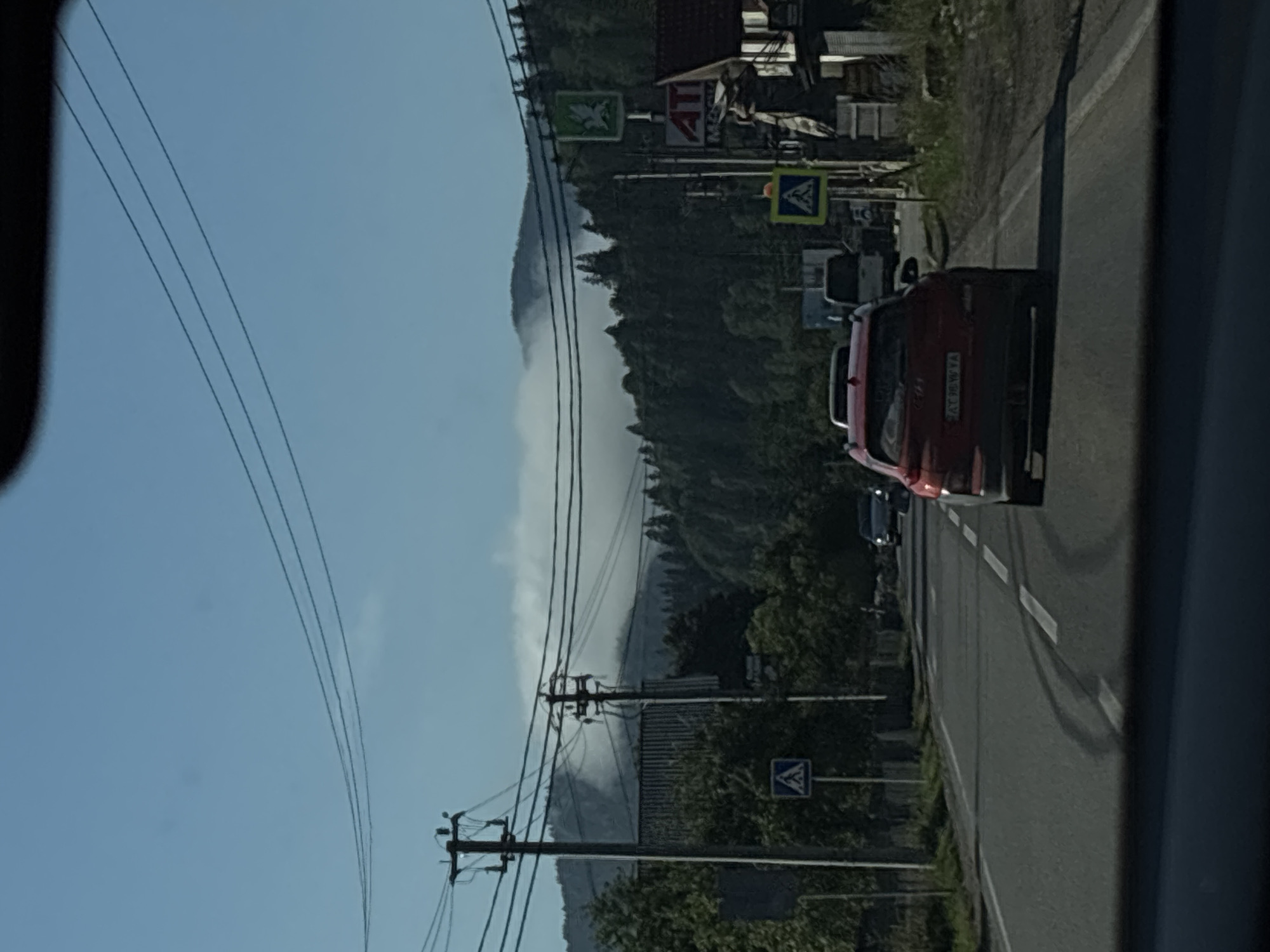 The image shows a street scene in Tatariv, Ukraine, with a red SUV in the foreground. The SUV is driving on a paved road with white lane markings. There are tire marks on the road, suggesting a recent sharp turn or braking. In the background, there are trees and a mountain range with fog or clouds obscuring the peaks. There are also power lines, utility poles, and buildings visible along the side of the road. One building has a sign with the Cyrillic letters "АТІ" and a green sign with a bird symbol. There are other vehicles on the road, including a white van. Pedestrian crossing signs are also visible. The sky is clear blue.