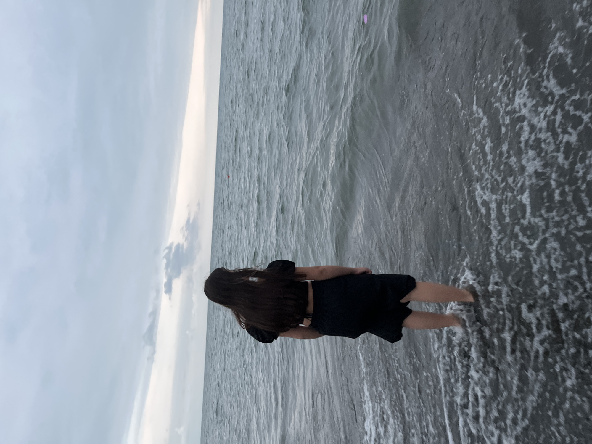 A young girl with long dark hair, wearing a black crop top and a black skirt, stands at the edge of the Black Sea in Ureki, Georgia. Her back is to the camera as she looks out at the choppy water. The sky is overcast with a soft, diffused light. There are two buoys visible in the distance, one red and one pink. The water is a dark gray-blue, and small waves are washing over the girl's feet. The shore is made of dark pebbles.
