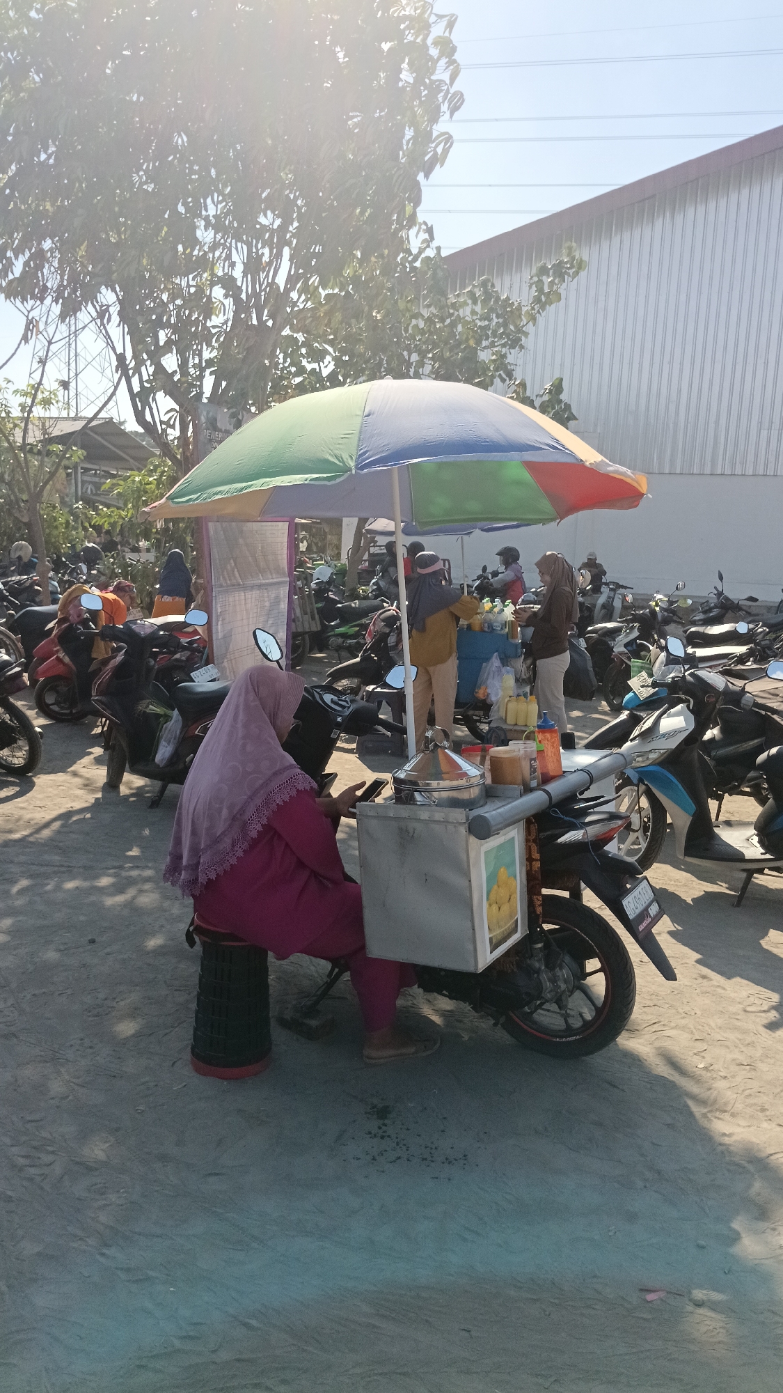 The image depicts an outdoor scene in what appears to be a marketplace or parking area, with numerous motorcycles parked in the background. A woman wearing a pink hijab and clothing is seated on a stool next to a motorbike that has a food cart attached to its rear. The cart is covered by a multi-colored umbrella. There are other individuals and stalls visible in the background, suggesting a commercial activity. The environment is sandy and sunlit, with trees and a large building with a corrugated metal exterior also present. The specific location is identified as Dukuh, Indonesia.