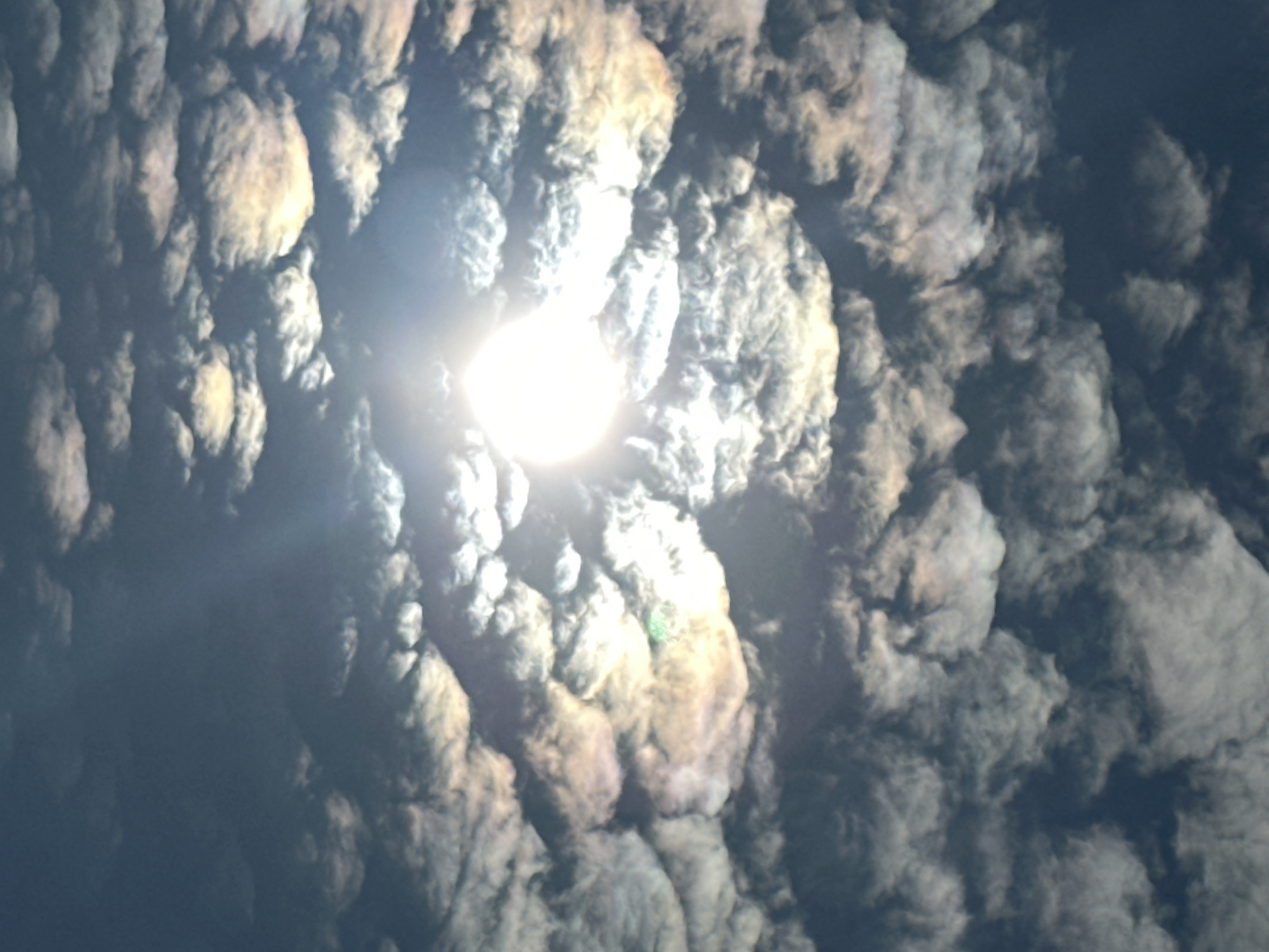 A sun, radiating intensely, is partially obscured by a thick blanket of clouds. The clouds exhibit iridescence, showcasing a spectrum of pastel colors including pinks, yellows, and greens, particularly around the sun. The sky itself is a deep blue, suggesting either a late afternoon or early morning light, or possibly a stormy weather pattern. The overall impression is one of dramatic natural lighting and atmospheric conditions. There are no people, buildings, or any other identifiable objects or actions present in this image. The location information provided for Podolsk, Russia, is not visually verifiable from the image content.