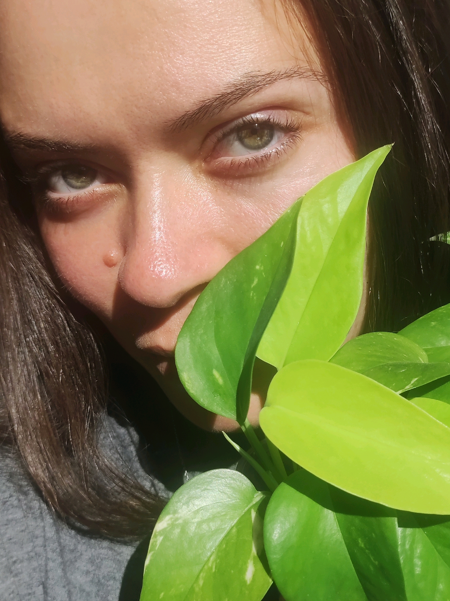 The image shows a close-up of a person's face, partially obscured by bright green leaves of a houseplant. The person's left eye, eyebrow, and part of their forehead are visible, with soft, natural light illuminating their skin. The eye is a light green color, and the skin appears smooth with some subtle texture. A small, light-colored mole is visible on the person's cheek. The background is dark and out of focus, with strands of dark hair framing the face. The overall impression is one of intimacy and natural beauty. The image is not geotagged, so its location in Chaikovsky, Russia, cannot be determined from the visual information alone.