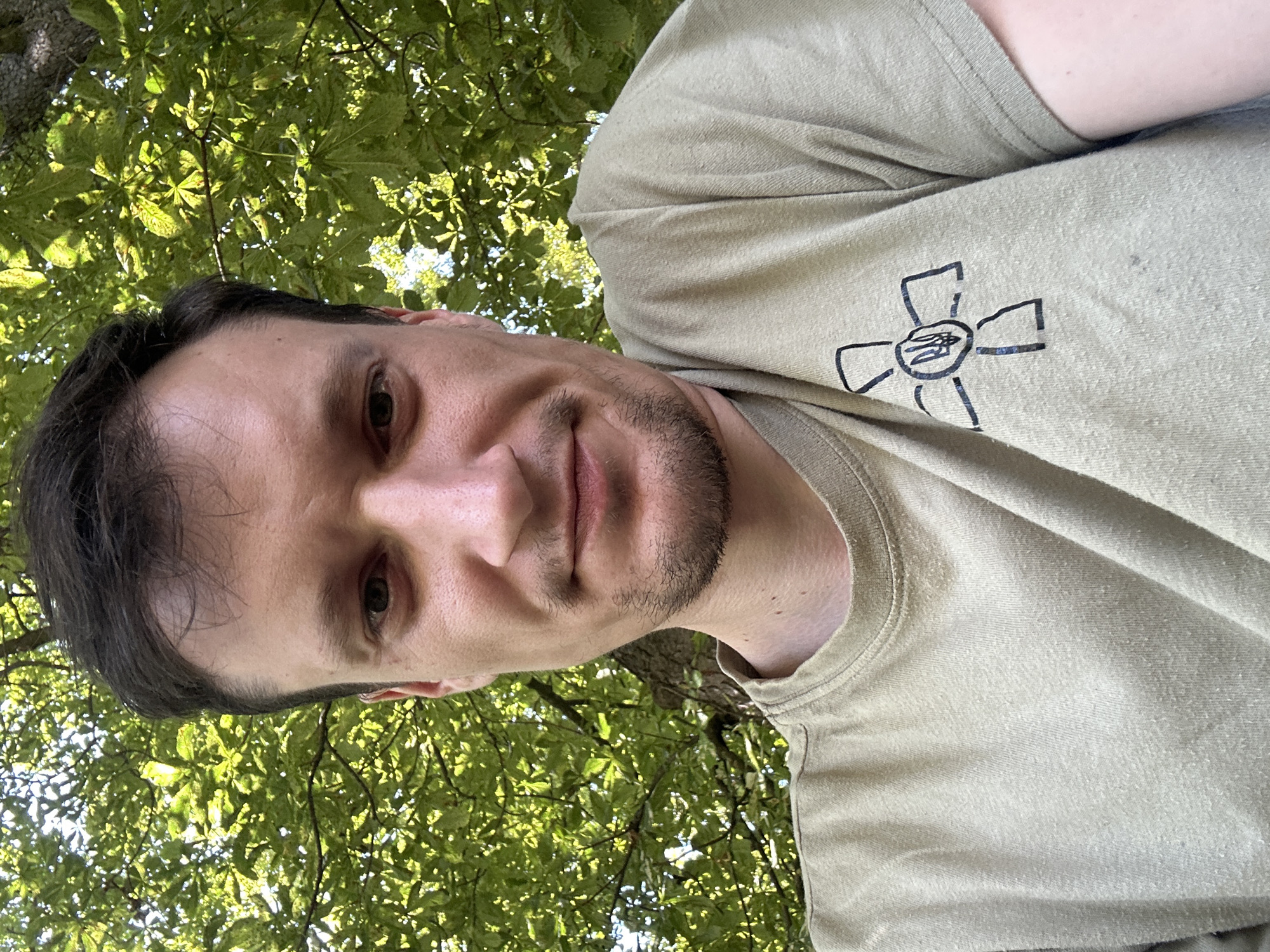 A man is taking a selfie outdoors, with green foliage from a tree in the background. He is wearing a tan t-shirt with a black symbol of a stylized cross with the Ukrainian trident in the center. The man has short dark hair and stubble on his chin. The scene appears to be in Makiv, Ukraine, based on the context provided.