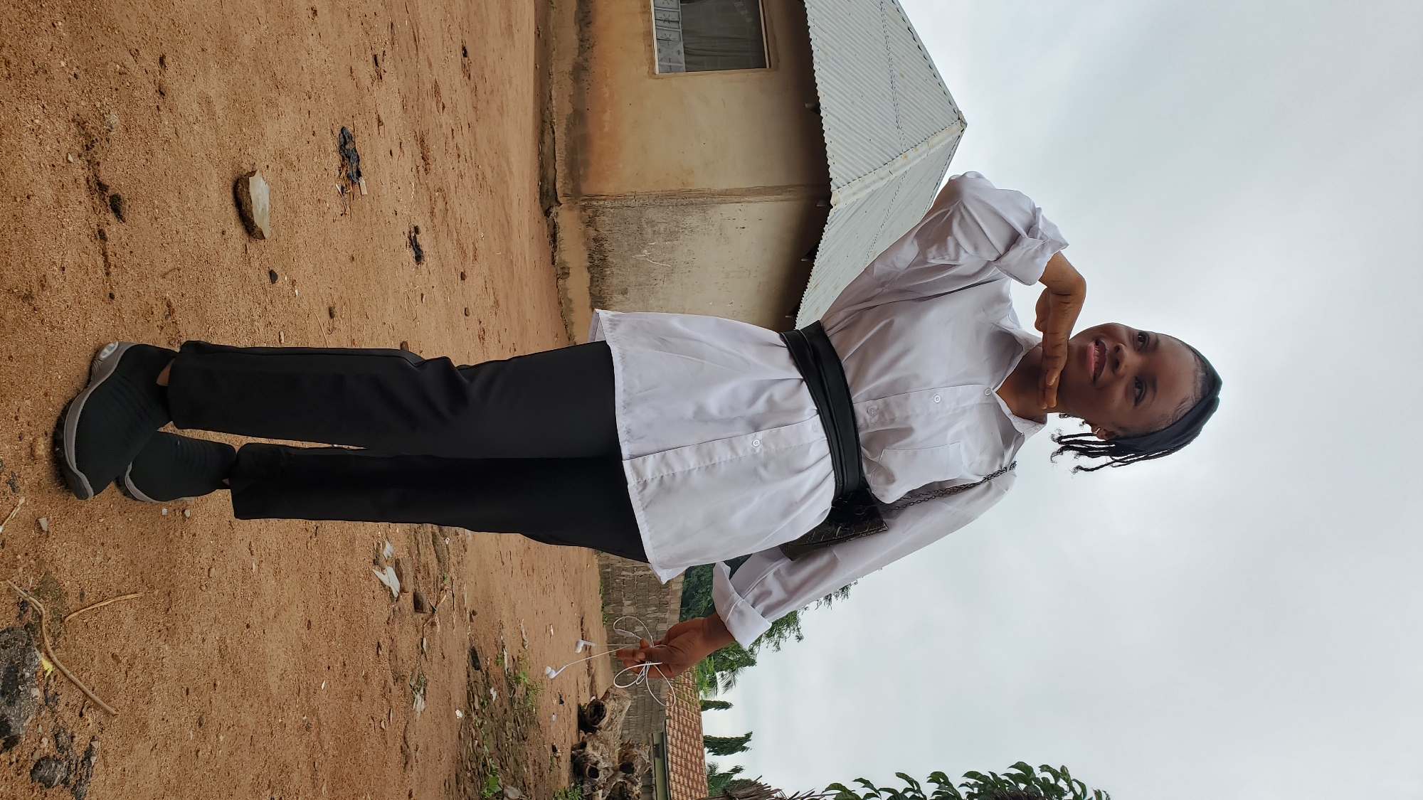 In Jalingo, Nigeria, a young Black woman stands outdoors on a dirt path. She is wearing a white button-down shirt, a wide black belt, and black pants. Her hair is styled in braids, and she is holding a pair of white earbuds in her right hand. A small brown purse with a chain strap is slung over her left shoulder. In the background, a small, weathered house with a corrugated metal roof is visible. The sky is overcast. The scene appears to be in a rural or semi-rural area.