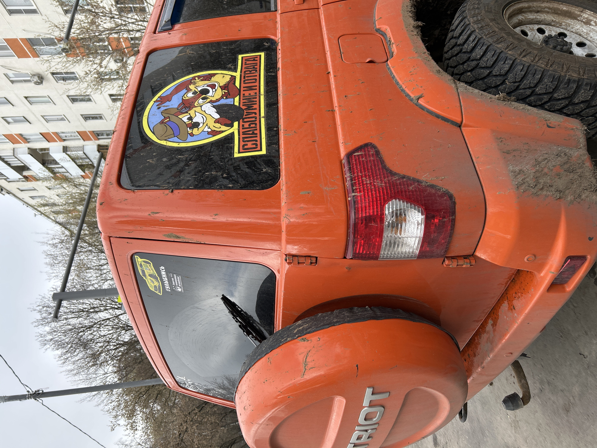 The image shows the rear of an orange UAZ Patriot SUV parked in Moscow, Russia. The vehicle is dirty, with mud splattered across its rear panels and spare tire cover. Stickers are affixed to the rear windows, including one depicting cartoon characters and a phrase in Russian: "СЛАБОУМИЕ И ОТВАГА" (Weakness and Courage). The spare tire cover reads "PATRIOT." The vehicle is parked near a building and trees, suggesting an urban environment. A power line is visible above the car.