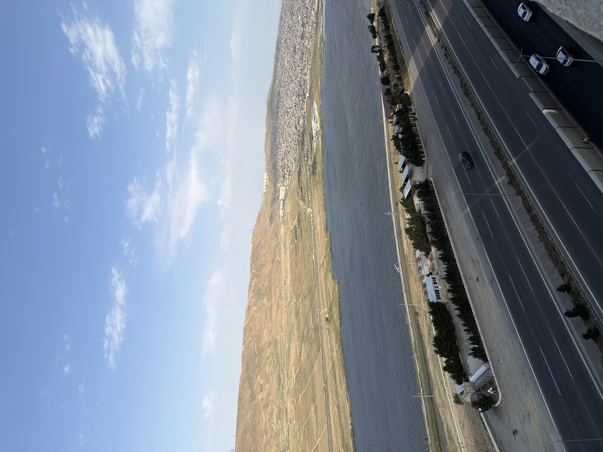 The image captures a wide-angle view of a coastal highway in Baku, Azerbaijan, adjacent to a body of water. The sky is a clear blue with scattered white clouds. In the distance, a densely populated city sprawls across a hillside, with numerous buildings. The foreground features a multi-lane highway with several vehicles in motion, including cars and what appears to be a truck. Along the side of the highway, there are rows of trees and some small structures, possibly rest stops or maintenance buildings. A large, dark body of water, likely the Caspian Sea, stretches along the left side of the highway, with a narrow strip of land separating it from the road. The land beyond the water is dry and hilly, with some signs of agriculture or cultivation in the form of patterned fields. The overall impression is one of urban development encroaching on a natural landscape.
