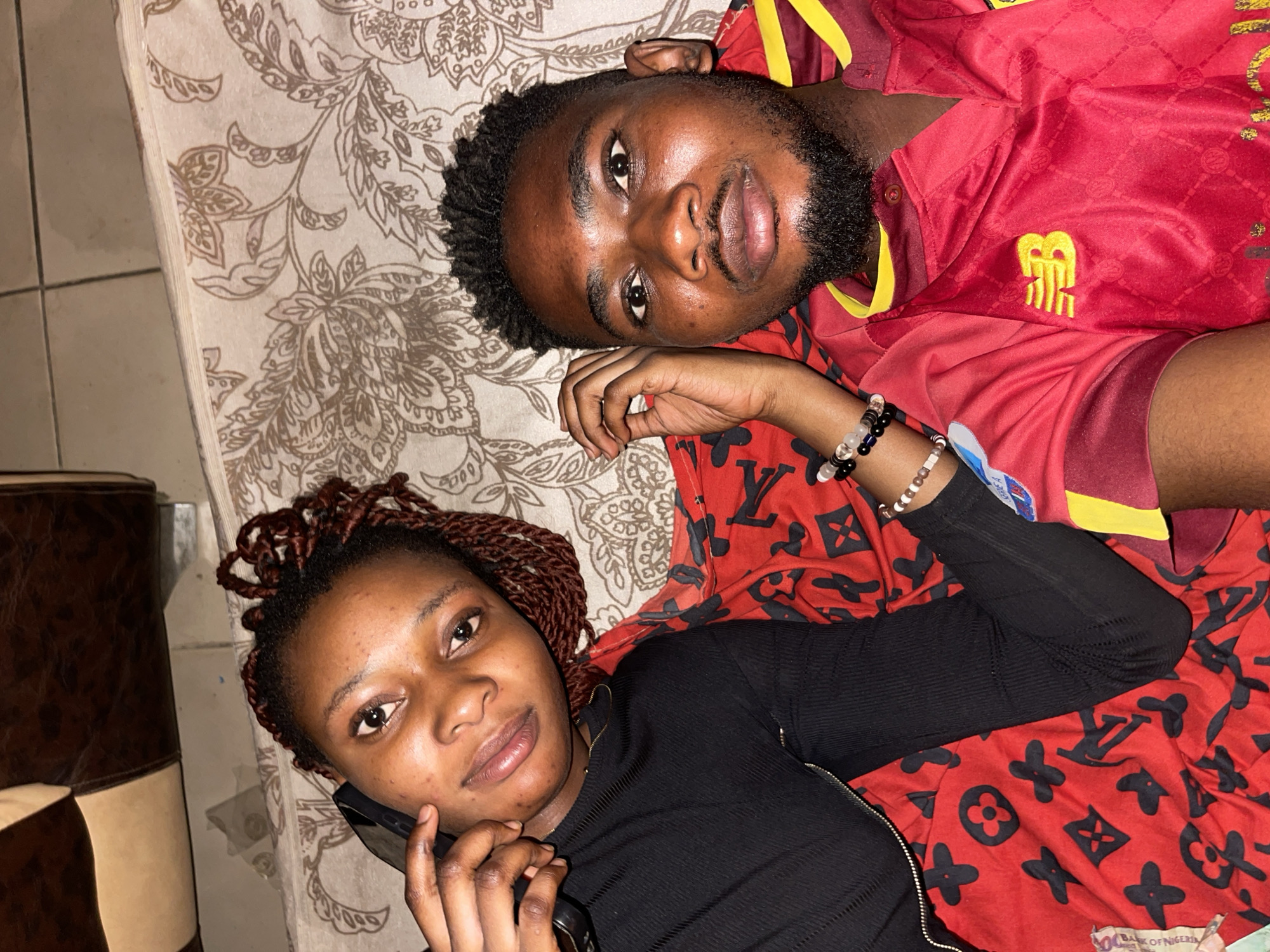 The image displays two young adults, a male and a female, lying down indoors in what appears to be a relaxed setting, likely in Unguwar Gabas, Nigeria.
The male, positioned on the right, has a short beard and dark, textured hair. He is wearing a red and yellow sports jersey adorned with a yellow emblem on the chest that resembles a stylized "BR" or similar logo. He has several beaded bracelets on his left wrist.
The female, on the left, has reddish-brown braided hair and is wearing a black long-sleeved ribbed top. She is holding a dark-colored smartphone to her ear, suggesting she is engaged in a phone call or listening to something. Beaded bracelets are visible on her right wrist.
They are lying on a surface covered with two distinct fabrics. The primary fabric underneath both individuals is a light-colored sheet with an elaborate brown floral pattern. Overlapping this, and covering the female, is a vibrant red fabric patterned with repeating black symbols that resemble Louis Vuitton monograms (flowers, circles, and 'LV' initials).
To the far left of the frame, a white tiled floor is visible, along with a brown and beige upholstered item, possibly a sofa armrest or chair. A partially visible currency note, showing the text "BANK OF NIGERIA," is tucked under the female's arm, providing a clear location cue for Nigeria. The indoor lighting suggests the photo was taken during the evening or night. Both individuals display calm or neutral expressions.