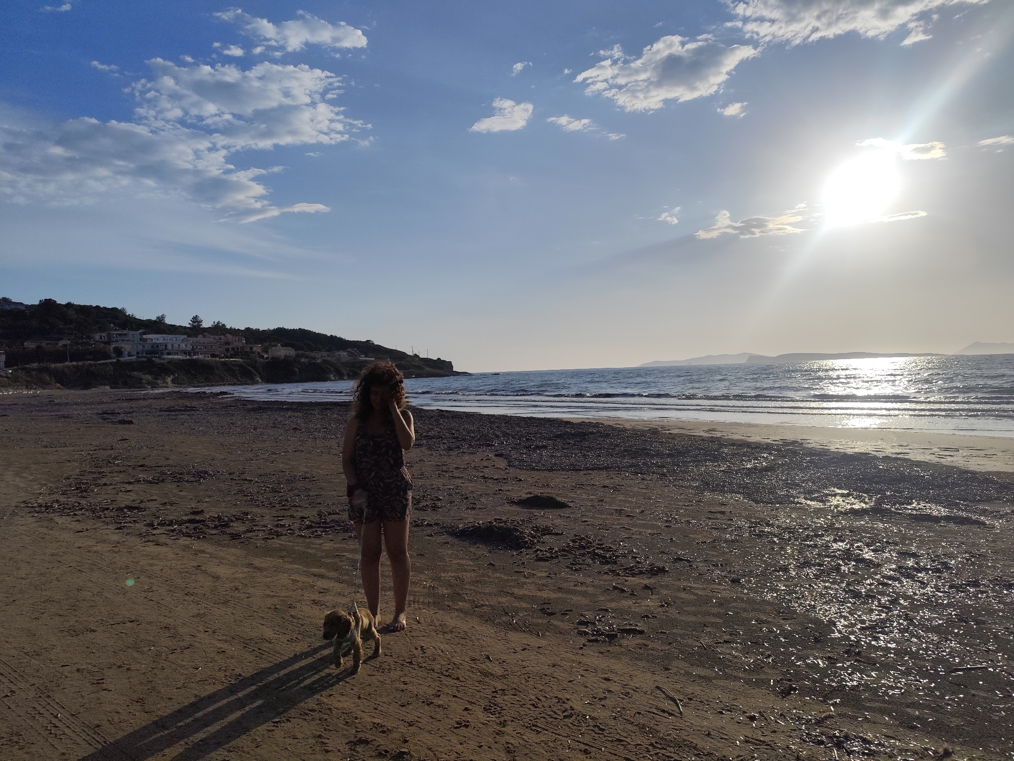 A woman and a small dog stand on a beach near Agios Stefanos, Greece. The woman is facing forward, her right hand near her face. She wears a patterned, knee-length dress and appears to be barefoot. A small brown dog is on a leash to her left. The beach is sandy, with some darker seaweed or debris scattered across it, and wet sand closer to the water. In the distance, there are buildings on a small hill, with water and land further out to the right. The sun is bright, creating a glare over the water. The sky is blue with some white clouds.