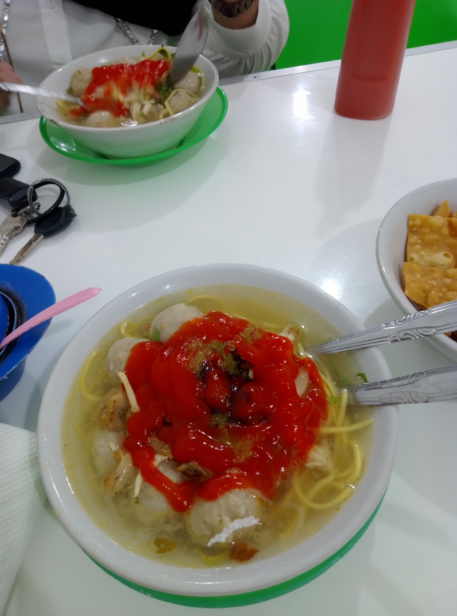 An overhead, close-up shot shows a table with food and personal items. In the foreground, a white bowl filled with broth, noodles, and meatballs is topped with a generous amount of red sauce and green garnishes. Two metal spoons rest on the rim of the bowl, with one partially submerged in the broth. To the right of the bowl, a second white bowl holds golden-brown fried wontons. The table itself is white and reflects light. In the background, a person wearing a white shirt is partially visible. To the left, a set of keys and a black wallet are on the table. A red bottle, possibly ketchup, stands upright in the background. The overall scene suggests a meal being enjoyed at a restaurant or a similar dining setting.
