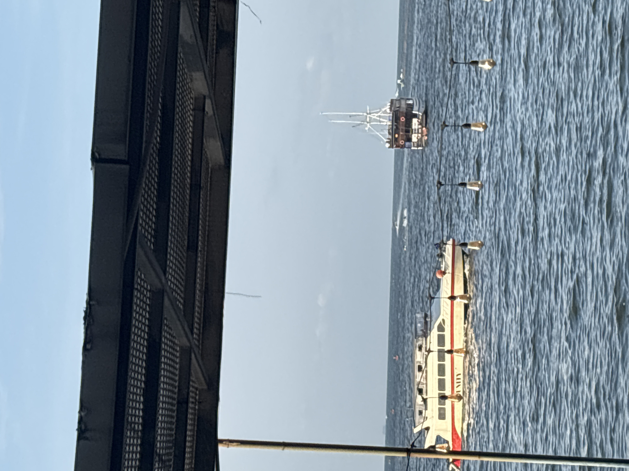 The image shows a view of the sea in North Jakarta, Indonesia, as seen from a structure with black metal grating on the left. In the sea, there is a white passenger boat with red accents and a sailing ship further in the distance. The water is rippled. There are several buoys or markers visible in the water. The sky is a pale blue color.