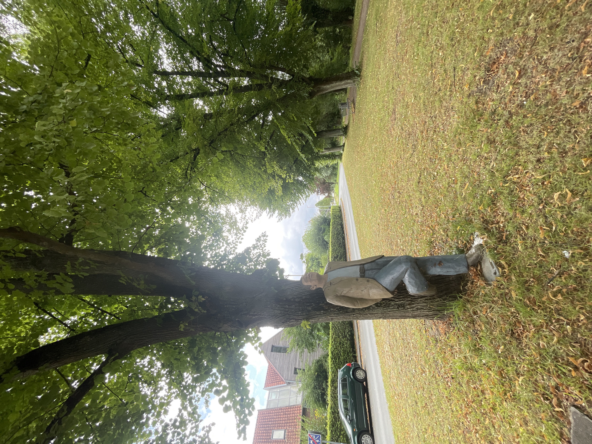 A statue of a man leaning against a tree is in the foreground of the image. The man is wearing a tan jacket, a light blue shirt, and blue jeans. He is looking up and to his left. The tree is large and has green leaves. The ground around the tree is covered in grass and fallen leaves. In the background, there is a road with a green car driving on it, a row of trees, and some buildings. The location is in Salzkotten, Germany.