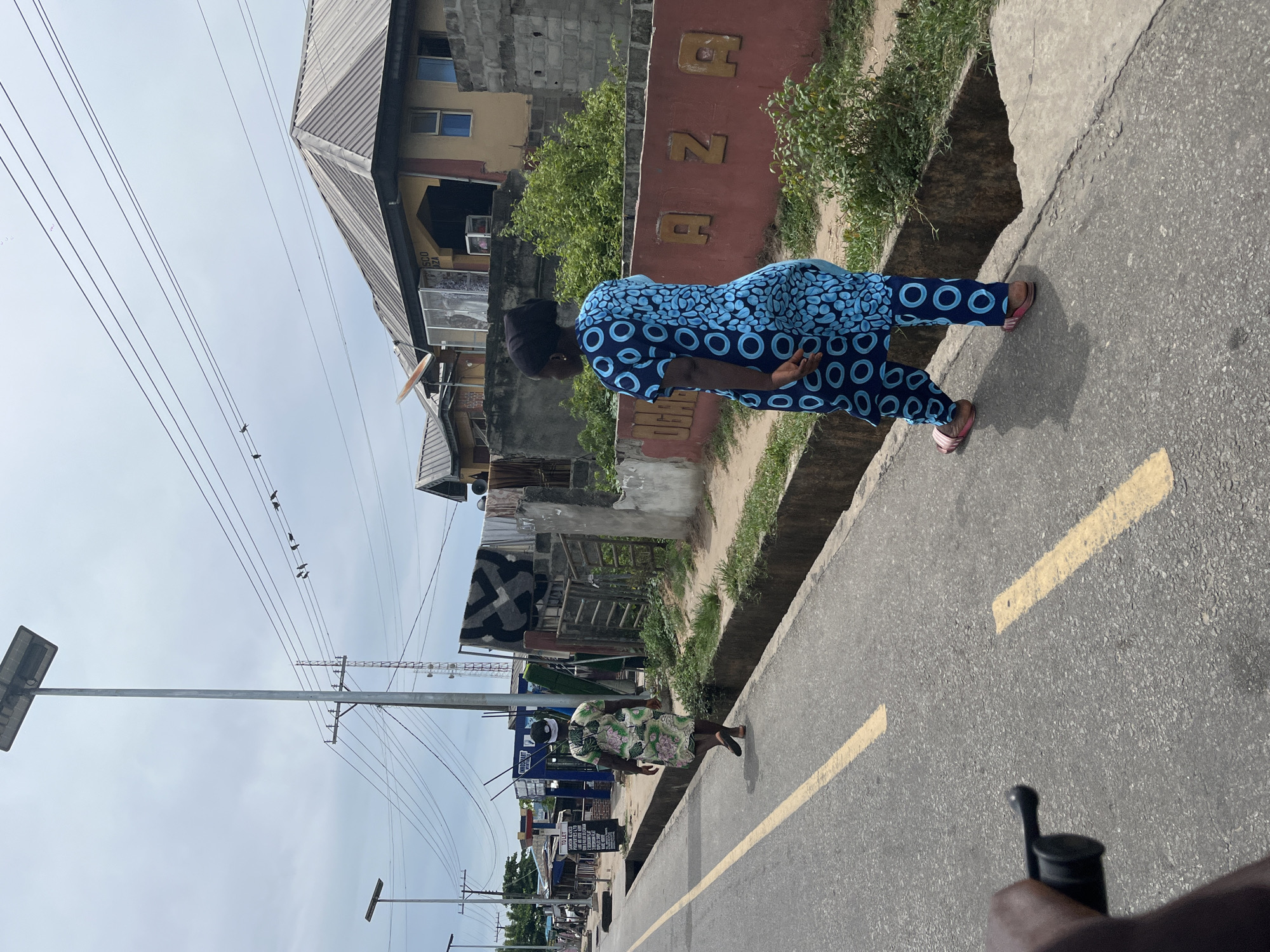 The image depicts a street scene in Badagry, Nigeria. A woman in a blue patterned jumpsuit walks along the side of a road with yellow road markings. The buildings and shops are varied, with structures including a yellow building with the red letters "AZA" on a wall and a structure covered with patterned materials. Power lines run overhead with birds perched on them. Another woman in a floral dress is walking in the same direction. The image is taken from a slightly low angle.
