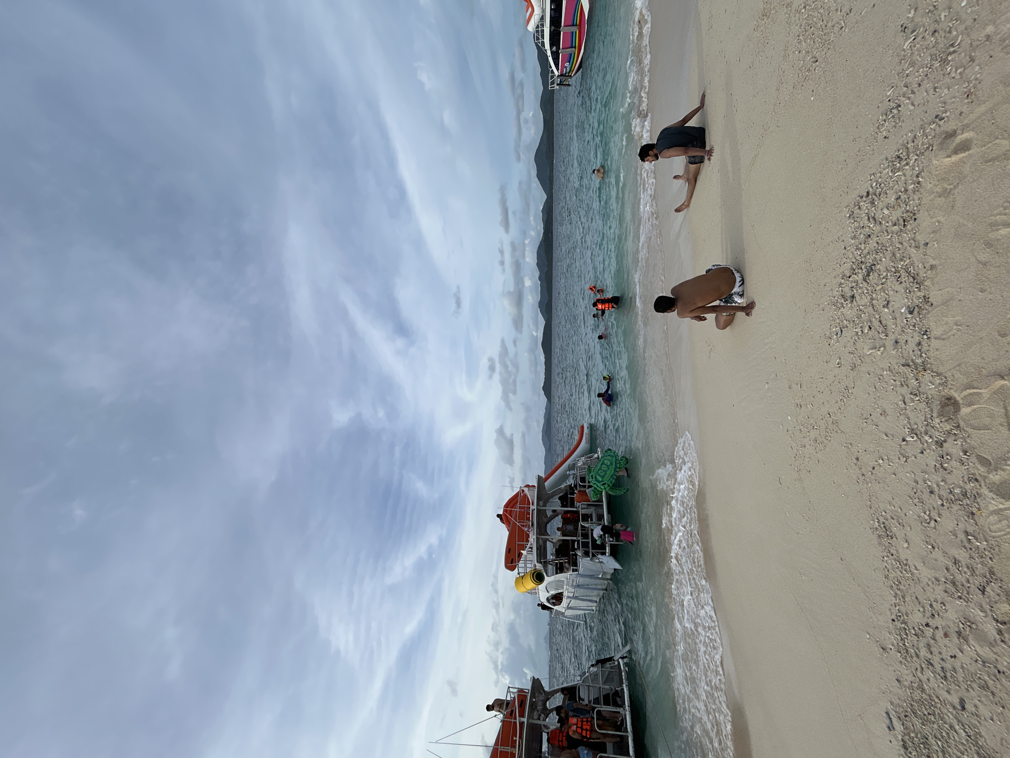 In Phru Nai, Thailand, two men sit on a sandy beach facing the ocean. The man on the left wears patterned swim trunks and faces away from the camera. The man on the right, wearing a dark tank top and shorts, faces left with his legs extended. In the background, several boats are anchored near the shore. One boat on the right has a colorful stripe along its side. Another boat on the left appears to be a tour boat with people on its deck. The water is clear and turquoise, with waves gently lapping the shore. Several people are visible swimming and wading in the ocean. The sky is overcast with scattered clouds. The beach is a mix of sand and small shells. The overall scene depicts a typical day at a beach resort.