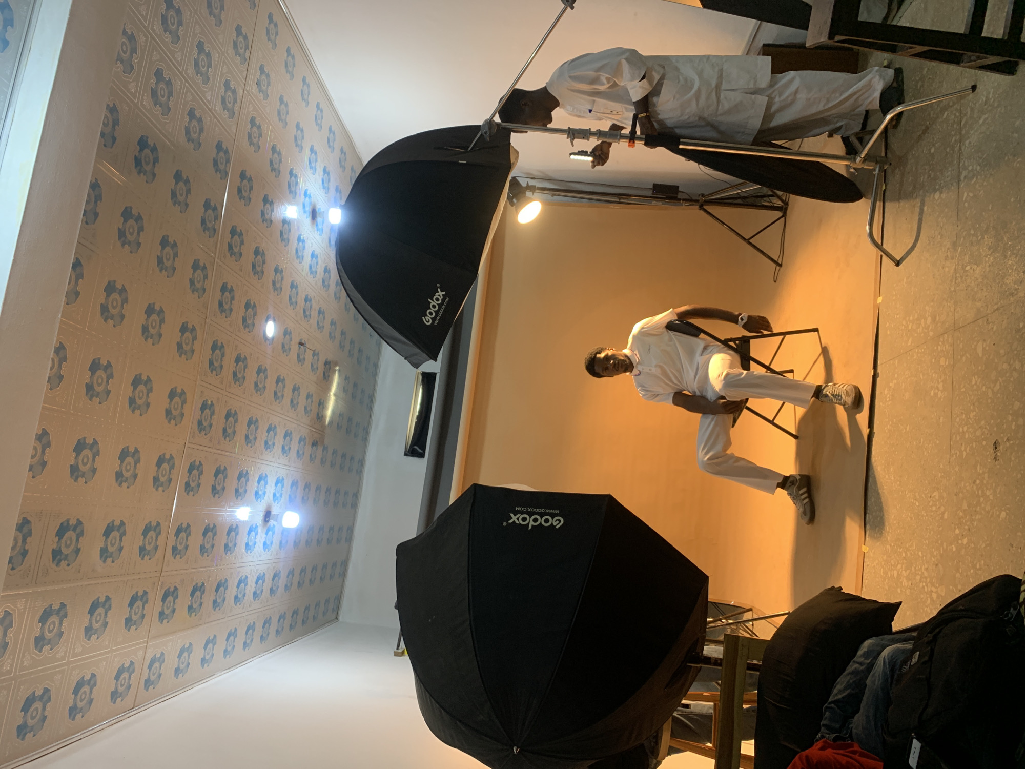 The photo captures a scene inside a room in Gura Top, Nigeria, seemingly set up for a photoshoot. A man in white is seated on a chair, illuminated by photography lighting equipment, including two large black Godox umbrellas. Another man, also in white, stands nearby adjusting the lighting. The room features patterned wallpaper on the ceiling and wall. There is additional equipment and clothing in the foreground.