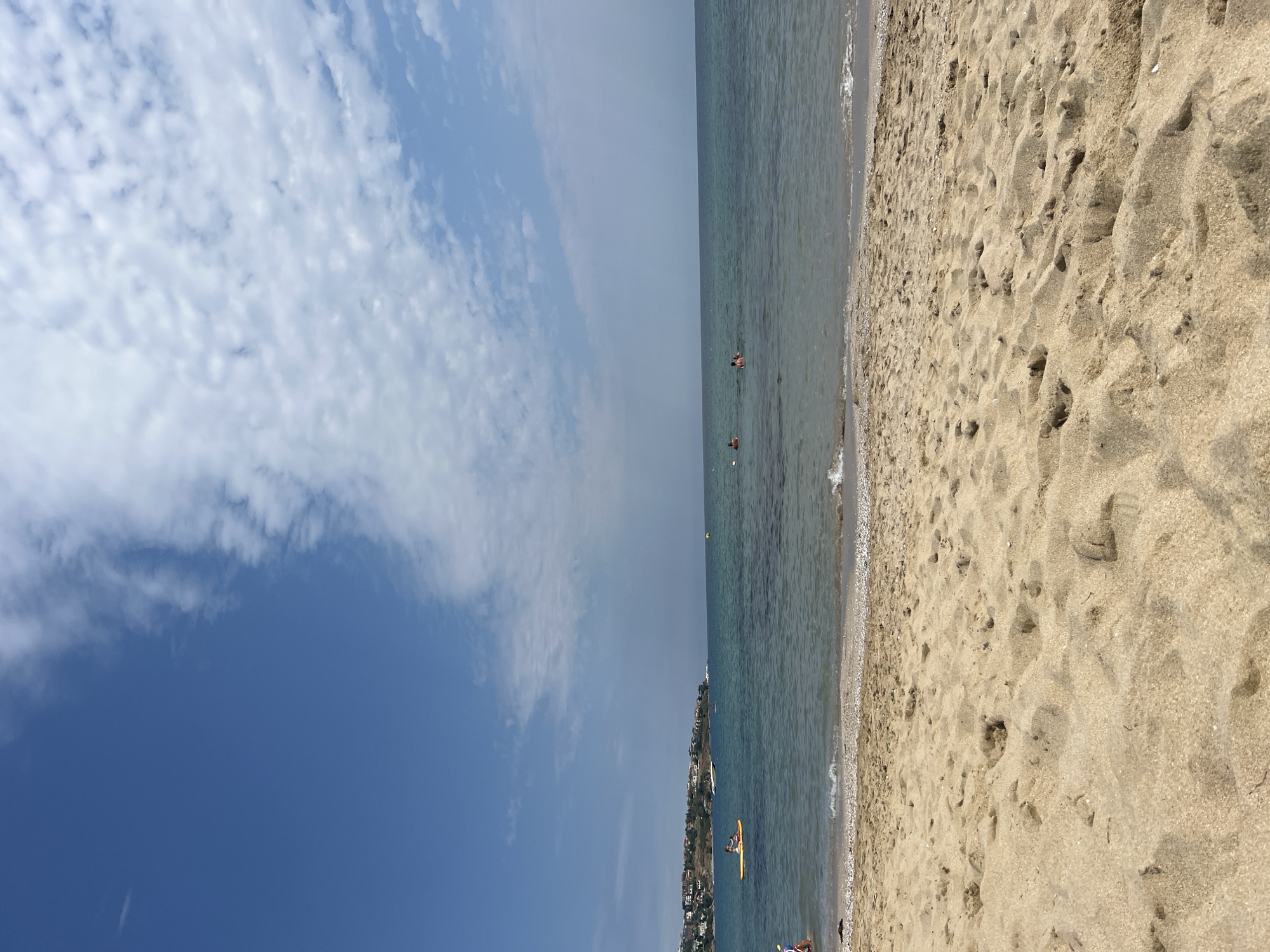 A tranquil beach scene unfolds under a vast blue sky dotted with scattered white clouds. The foreground is dominated by a sandy beach, showing footprints and scattered shells, with gentle waves lapping at the shore. In the middle ground, the calm, clear blue sea stretches towards a distant coastline. A cluster of buildings, appearing to be houses with red roofs, is visible on this coastline, suggesting the location is near the town of Sozopol, Bulgaria. Several people are enjoying the water: one person is on a yellow paddleboard, and a few others are swimming or wading. Two small boats are also present in the distance, adding to the peaceful atmosphere. The overall impression is one of a relaxed summer day at the seaside.