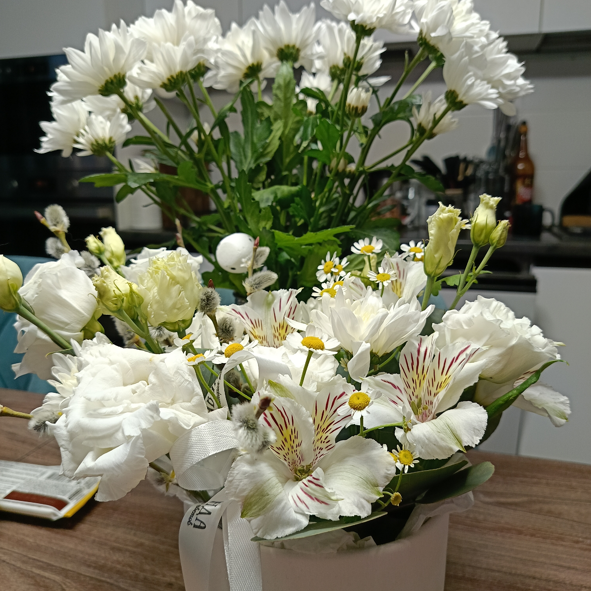 Two bouquets of white flowers sit on a wooden table. The first bouquet is composed of white chrysanthemums. The second bouquet is composed of white roses, alstroemeria with red stripes, daisies, and pussy willow. The second bouquet is tied with a white ribbon and sits in a white container. A box sits on the table next to the bouquet. The location appears to be a kitchen in Markovskii, Russia.