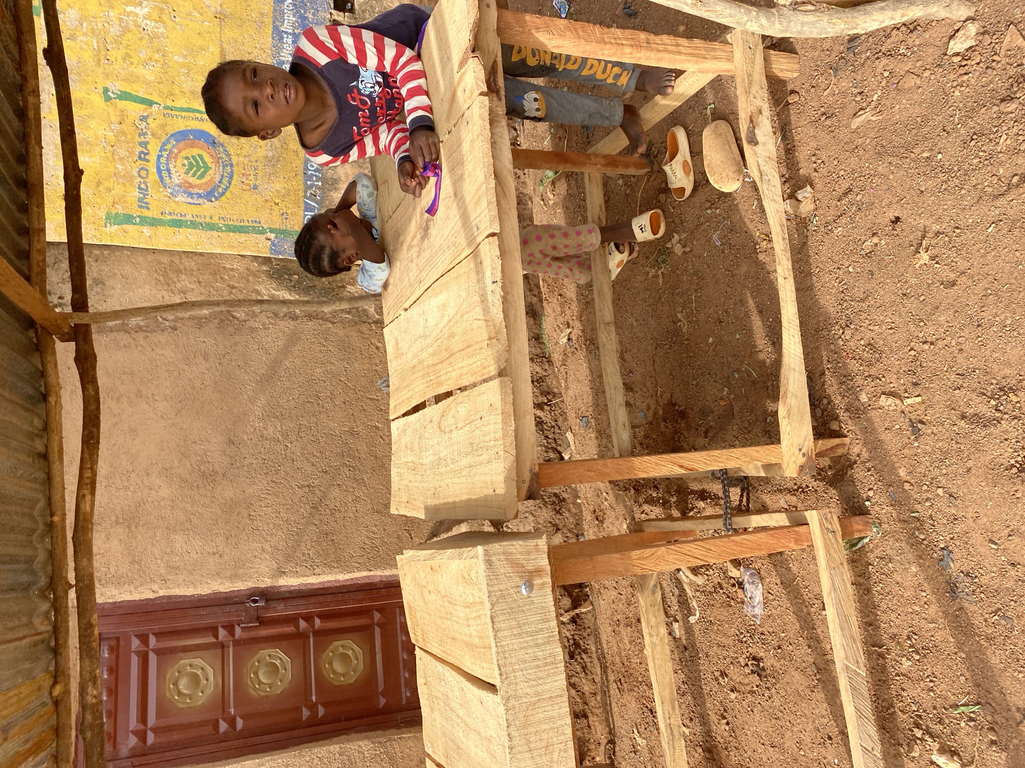 Here is a detailed description of the image:
**Location:** Bukuru, Nigeria
**Scene:** The image captures an exterior scene next to a building. The building has a brown door with a symmetrical design of round inlays and a yellow/tan wall next to the door, with yellow burlap next to that. Adjacent to the building is a partially constructed wooden structure, possibly a piece of furniture or a building frame. The ground is dirt, with some debris such as plastic trash scattered around.
**People:** Two children are present in the image. One is wearing a red and white striped shirt with a cartoon character on it. The other child is wearing a light blue shirt. The two children are standing near the wooden structure. Two more children can be seen underneath the wooden structure.
**Objects:**
* Wooden structure (partially constructed)
* Building with door and wall
* Yellow burlap
* Sandals
* Debris/trash on the ground
* Plastic ribbon
**Action/Interactions:** The children appear to be interacting with the wooden structure. One child is holding a piece of plastic ribbon.