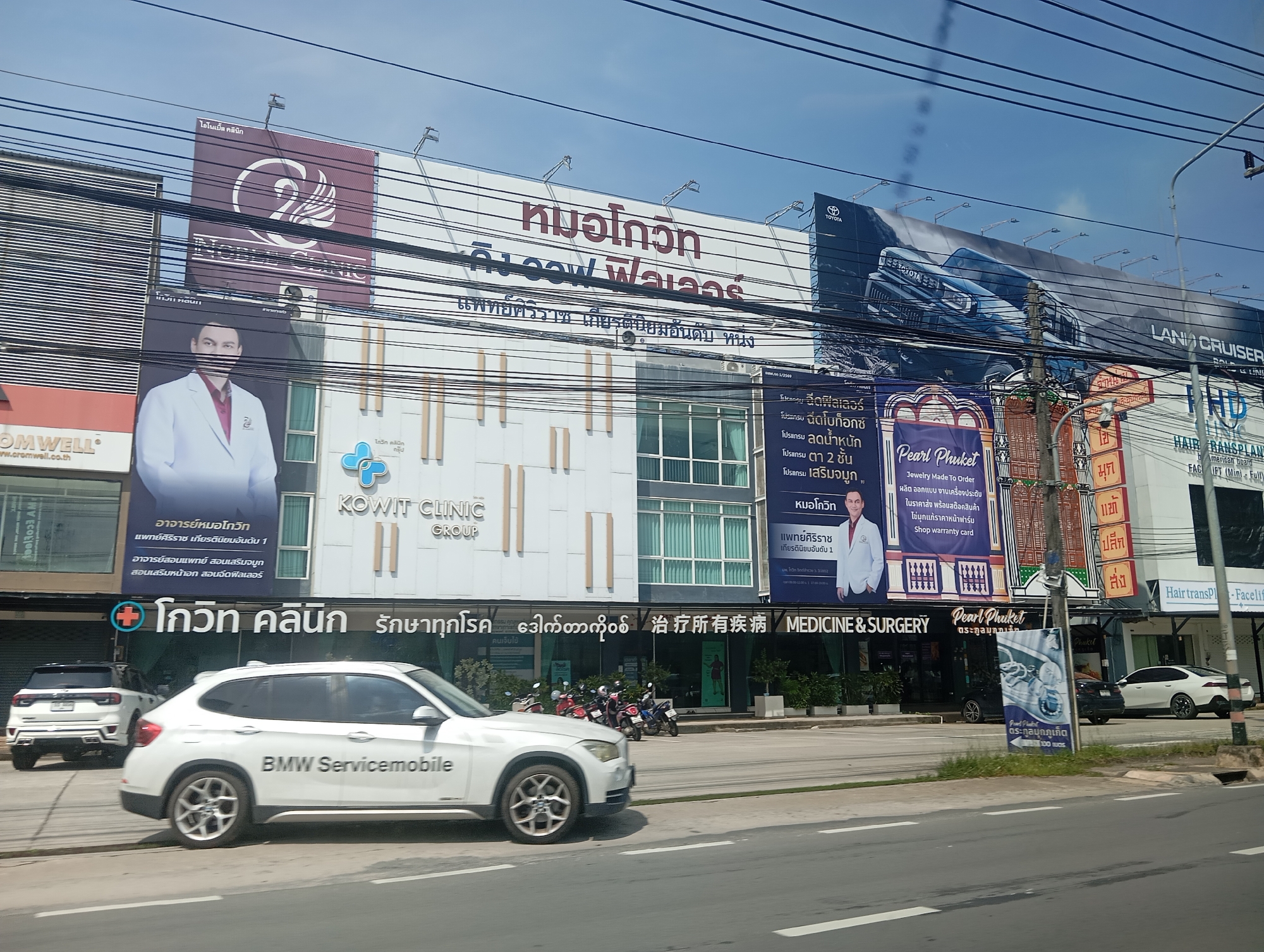 In Ko Kaeo, Thailand, a street scene unfolds with numerous billboards advertising various businesses. A prominent billboard features a white swan logo and the text "MONOCLINIC". Next to it, another billboard displays the words "KOWIT CLINIC GROUP". Further along, a Toyota Land Cruiser advertisement stretches across a large billboard. Below these, smaller signs advertise services such as "Pearl Phuket" and "Hair transPL".
In the foreground, a white BMW Servicemobile is parked on the side of the road, with another white SUV visible behind it. Several motorcycles are also parked near the buildings. The buildings themselves appear to be shops or clinics, with large windows and modern facades. The sky is clear and blue, indicating a sunny day. The overall scene depicts a busy commercial area with a mix of advertising and vehicle traffic.