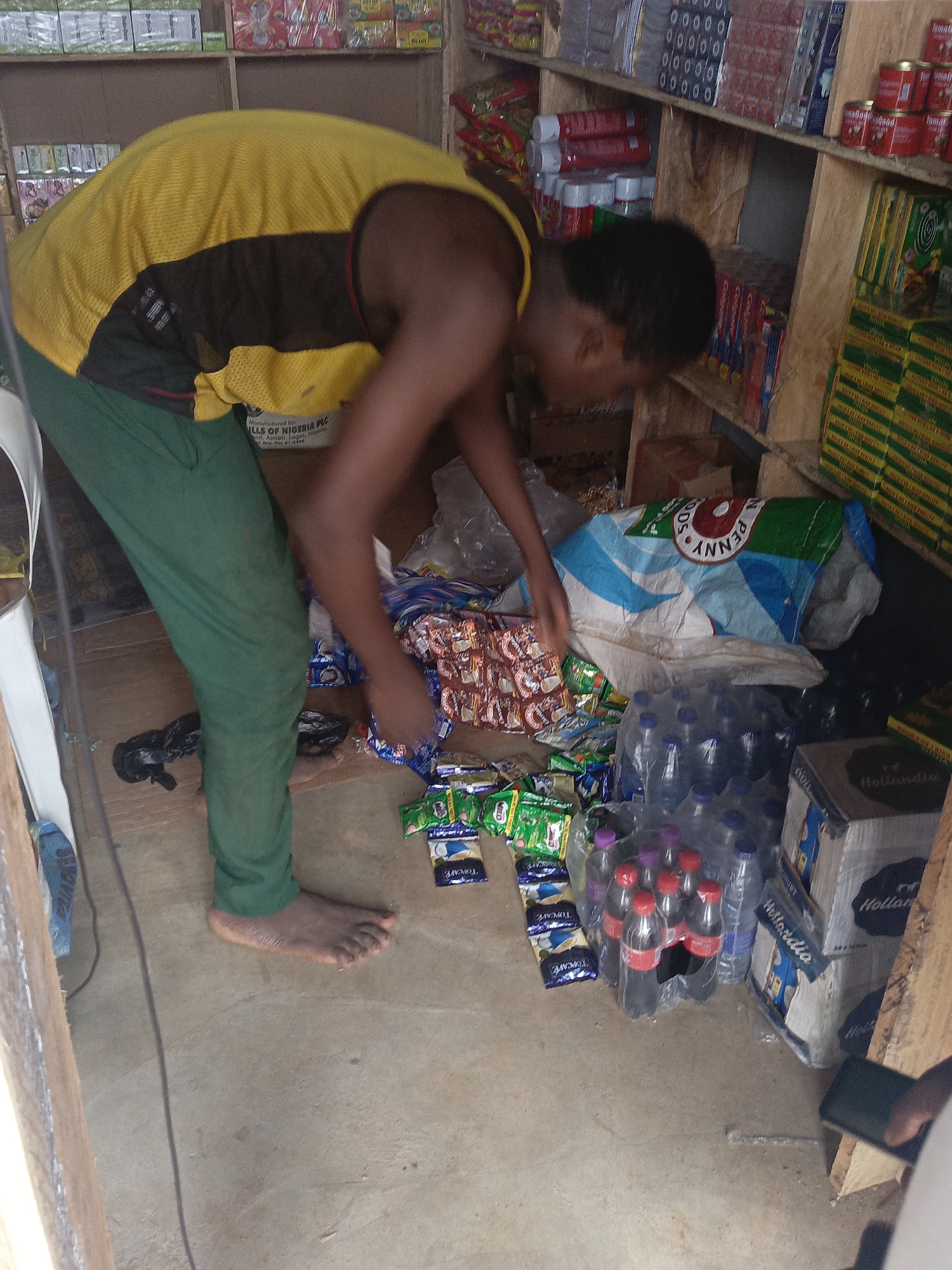A young man with bare feet stands in a small, cluttered shop in Akwanga, Nigeria, bending over to sort through goods on the floor. He wears green pants and a yellow and black tank top. In front of him, there are various items scattered on the floor, including bottled water, packets of biscuits, and bags of what appears to be food products. Some of the brand names visible are "Penny" and "Hollandia". Shelves laden with goods can be seen in the background. The environment appears to be a small, local store in Akwanga, Nigeria.