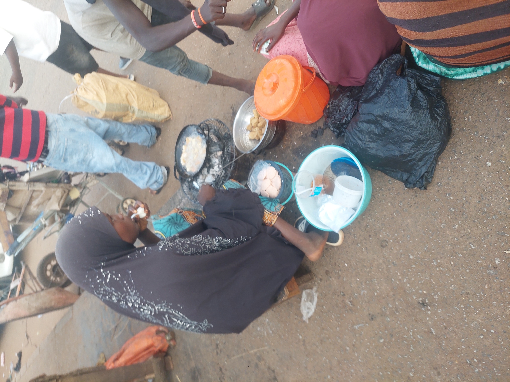The image depicts an outdoor street food vending scene within an urban setting. In the foreground, a woman, wearing a dark head covering with white patterns and a blue-patterned dress, sits on a low wooden stool. She is actively engaged in food preparation, with her right hand holding a small white object near her mouth. In front of her, a black frying pan containing light-colored food items rests on a small metal grill over embers. To her right, a metal colander holds additional fried food. Surrounding her workstation on the ground are several plastic containers: a dark blue bucket holds flat, round, light-colored food items sealed in a clear plastic bag, an orange lidded bucket, and a light blue bowl containing various plastic cups and containers. A black plastic trash bag is situated near the right side of the frame.
Several other individuals are present. To the left and slightly behind the vendor, a man wearing a red and black striped shirt and light blue jeans stands, facing away. Further back, another man in a white t-shirt and dark shorts stands, observing the scene. To the right of the vendor, a person wearing a light brown shirt and orange wristbands is partially visible, looking down. On the far right, the lower body of another person, clad in a pink garment and a brown and black striped garment, is visible, with a hand resting on the pink fabric, holding a small white item.
The ground is a rough, dirty surface composed of concrete and gravel with scattered debris. In the background, partially visible elements include a wheelbarrow or cart and indistinct structures, characteristic of a street market or roadside location in the city of Unknown, Unknown.