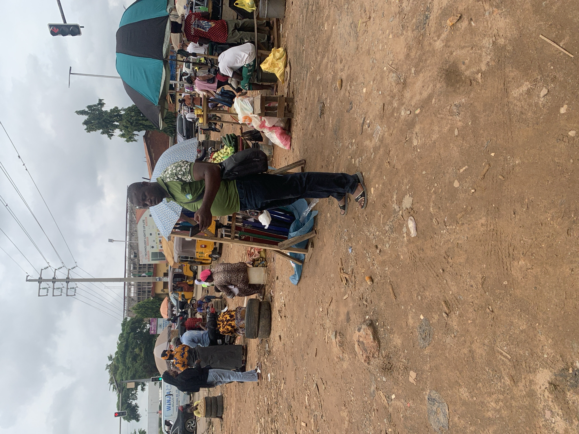The image depicts a busy outdoor market scene in Abuja, Nigeria, under an overcast sky. In the foreground, a man wearing a green and camouflage-patterned shirt and dark pants stands with a black bag slung over his shoulder, looking at his phone. The ground is dirt and appears worn.
Further into the scene, several people are present in the market. To the left, three individuals, two women and one man, are gathered near a stack of tires. One woman wears a black top and jeans, while another is in a vibrant yellow and orange patterned dress with a grey skirt. The man is in a light blue shirt. In the middle ground, a woman in a leopard-print outfit sits on a bucket, facing away from the camera. Behind her, two stacked tires are visible.
Various market stalls are set up with umbrellas providing shade. Yellow and black umbrellas are prominent, as well as a blue and green striped umbrella. Tricycles, commonly used as taxis in Nigeria, are parked in the background, suggesting transportation is readily available. People are seen interacting around these stalls, some appearing to be vendors and others customers. Bags of goods, including what appear to be sacks of rice and produce, are visible. In the far background, buildings and power lines are present, indicating an urban environment. Traffic lights and a road suggest this market is located near a busy intersection or street. The overall impression is one of a lively and active marketplace in Nigeria's capital city.