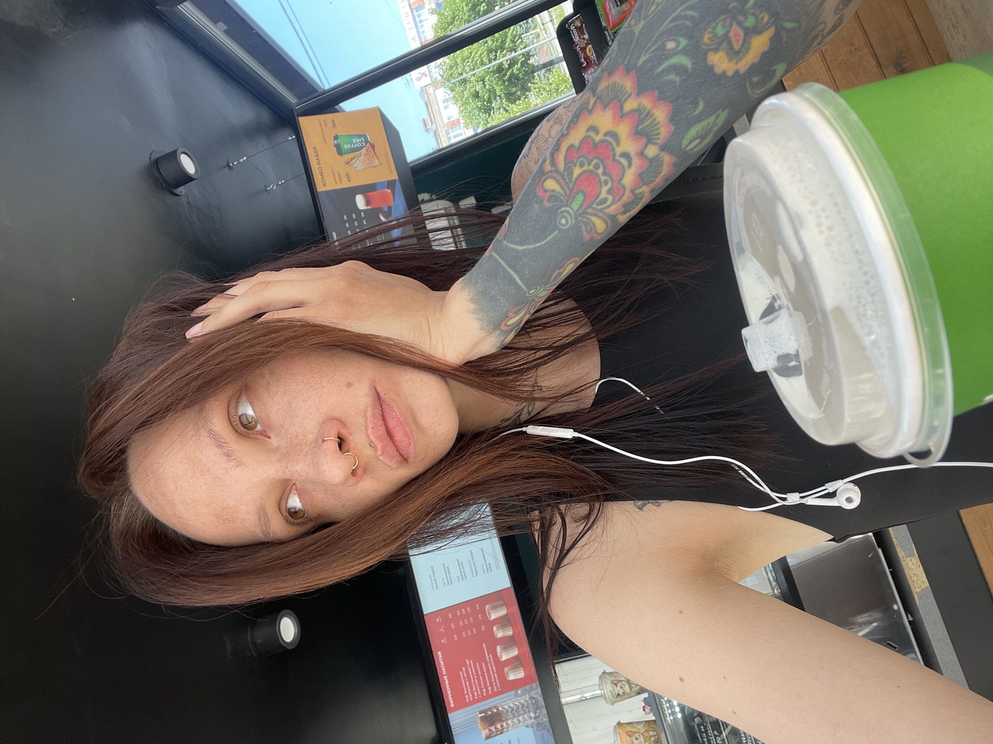 A person with auburn hair, light skin, and a tattooed arm is in a cafe. The person has a piercing in their nose and is wearing earphones. A green cup with a lid is on the table beside them. The cafe has a black counter and a menu board with Russian text. Through the window, trees and buildings are visible, suggesting an urban setting. The location is Izhevsk, Russia.