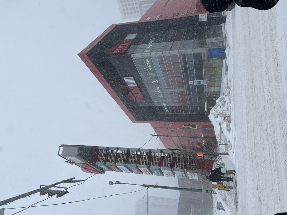 A multi-story building with a distinctive red trim is featured in snowy weather. Signage and glass windows adorn the building's facade. A tall, elongated structure with multiple billboards or signs is visible in the foreground. Snow covers the ground and street, indicating winter conditions. Pedestrians are seen on the snowy sidewalk. Traffic signals and overhead wires are also present in the wintry urban scene.
