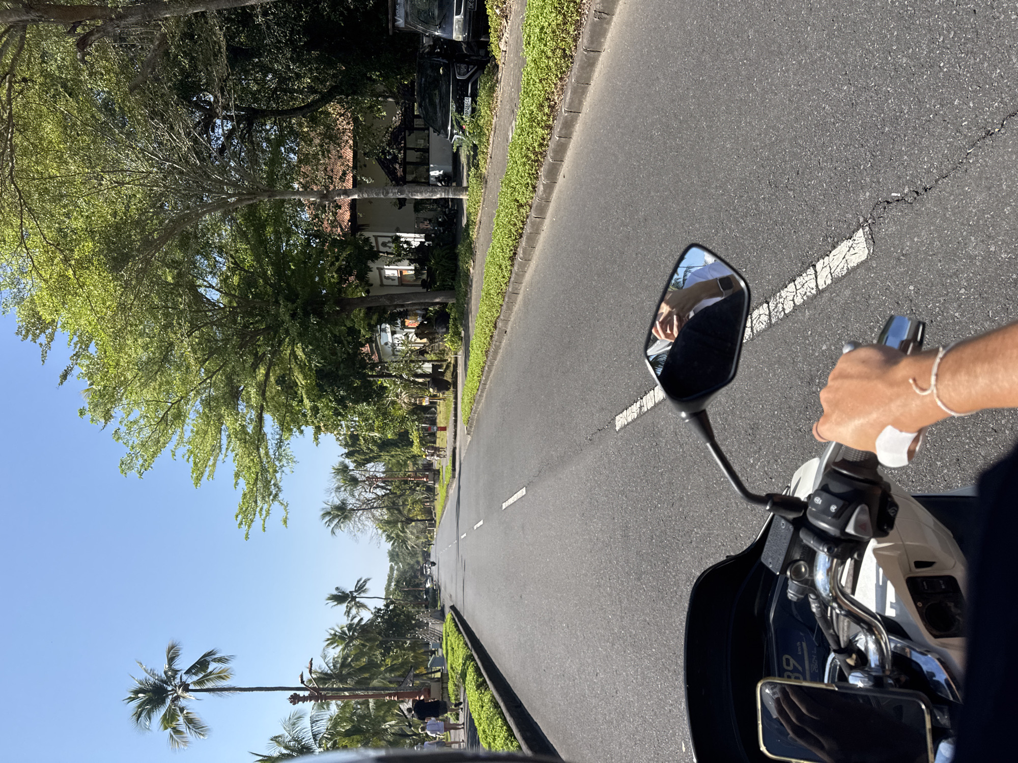 The image shows a point-of-view shot from someone riding a scooter on a paved road. The road is lined with lush green vegetation and palm trees on both sides, suggesting a tropical or subtropical location. On the left side of the road, several people are walking, and there is a small wall with manicured bushes. On the right side, there are buildings, some with terracotta roofs, and parked vehicles, including a black SUV and a black van. The sky is clear and blue, indicating a sunny day. The scooter's dashboard is partially visible, showing a speed of 39 km/h. A hand is visible on the right handlebar, wearing a bracelet. The scooter's mirror reflects the rider's face and arm. The overall scene depicts a leisurely ride along a tree-lined street in a town or resort area.