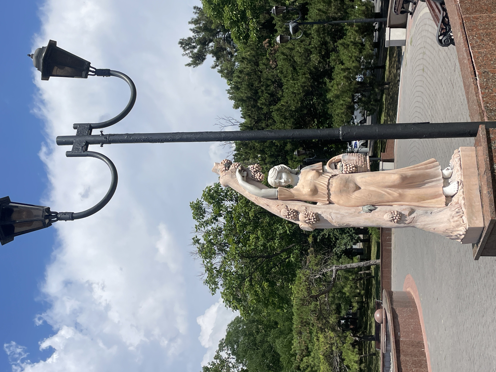 The image shows a statue of a woman holding grapes in a park setting. The statue is made of a light-colored material, possibly marble or stucco, and depicts a woman in a flowing dress with a basket of grapes. She is standing next to a tree trunk that is also adorned with grapes. A double lamppost with two lanterns stands behind the statue. In the background, there are lush green trees, a paved walkway with a circular pattern, benches, and a low-lying fountain or decorative wall. The sky is blue with scattered white clouds. This scene is located in Novorossiysk, Russia.