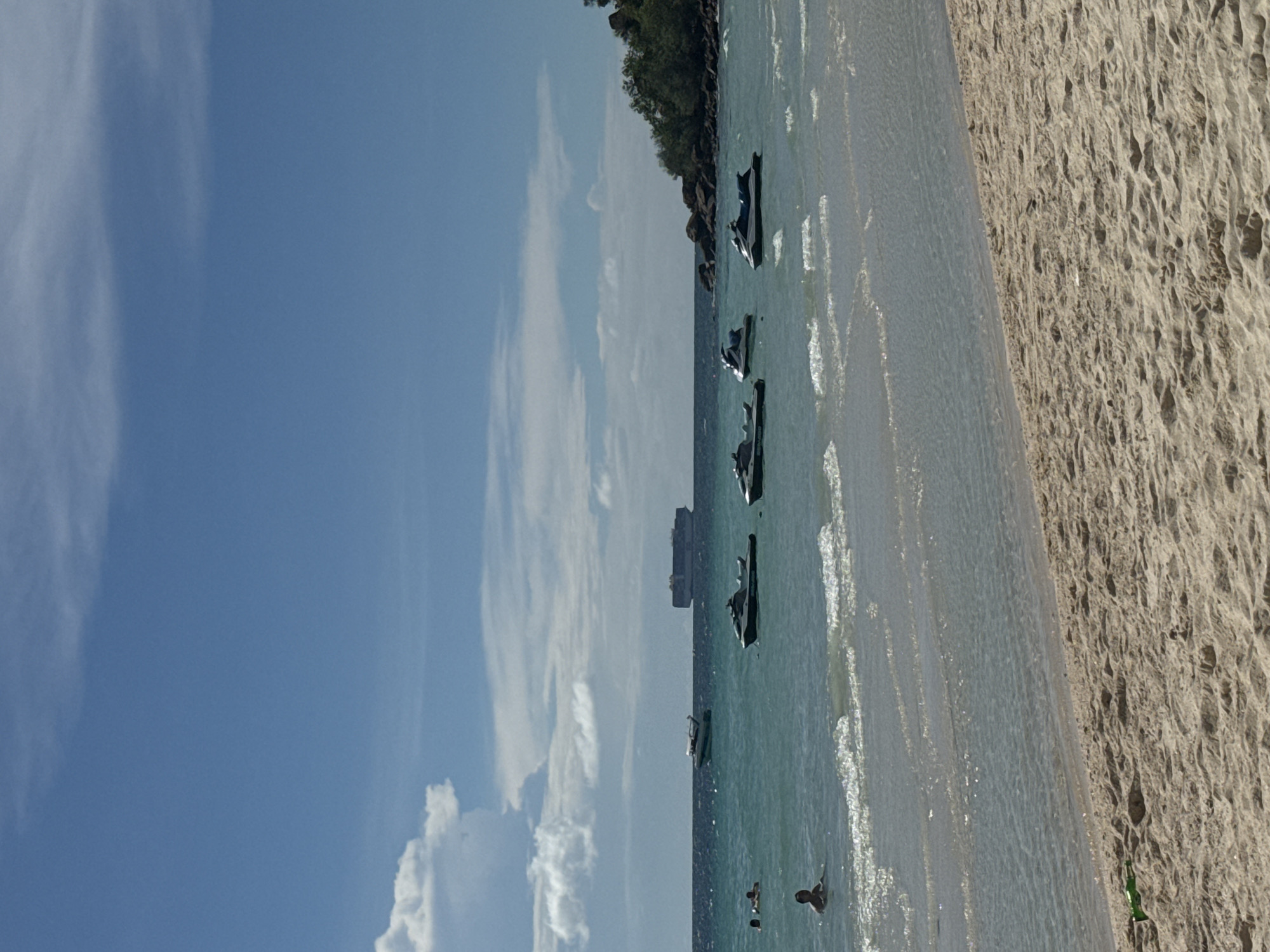 This image shows a beach scene, possibly in Pattaya City, Thailand. The perspective is from the shore looking out towards the sea. The scene includes a sandy beach on the right side of the frame and a clear blue sky with scattered white clouds to the left. The water has various shades of blue and white due to the waves. Several jet skis are parked along the coastline, with a few boats further out in the sea. Near the beach, there are a few people swimming or standing in the water. The shoreline curves into the distance and is lined with greenery, which could be vegetation or trees growing near the coast.