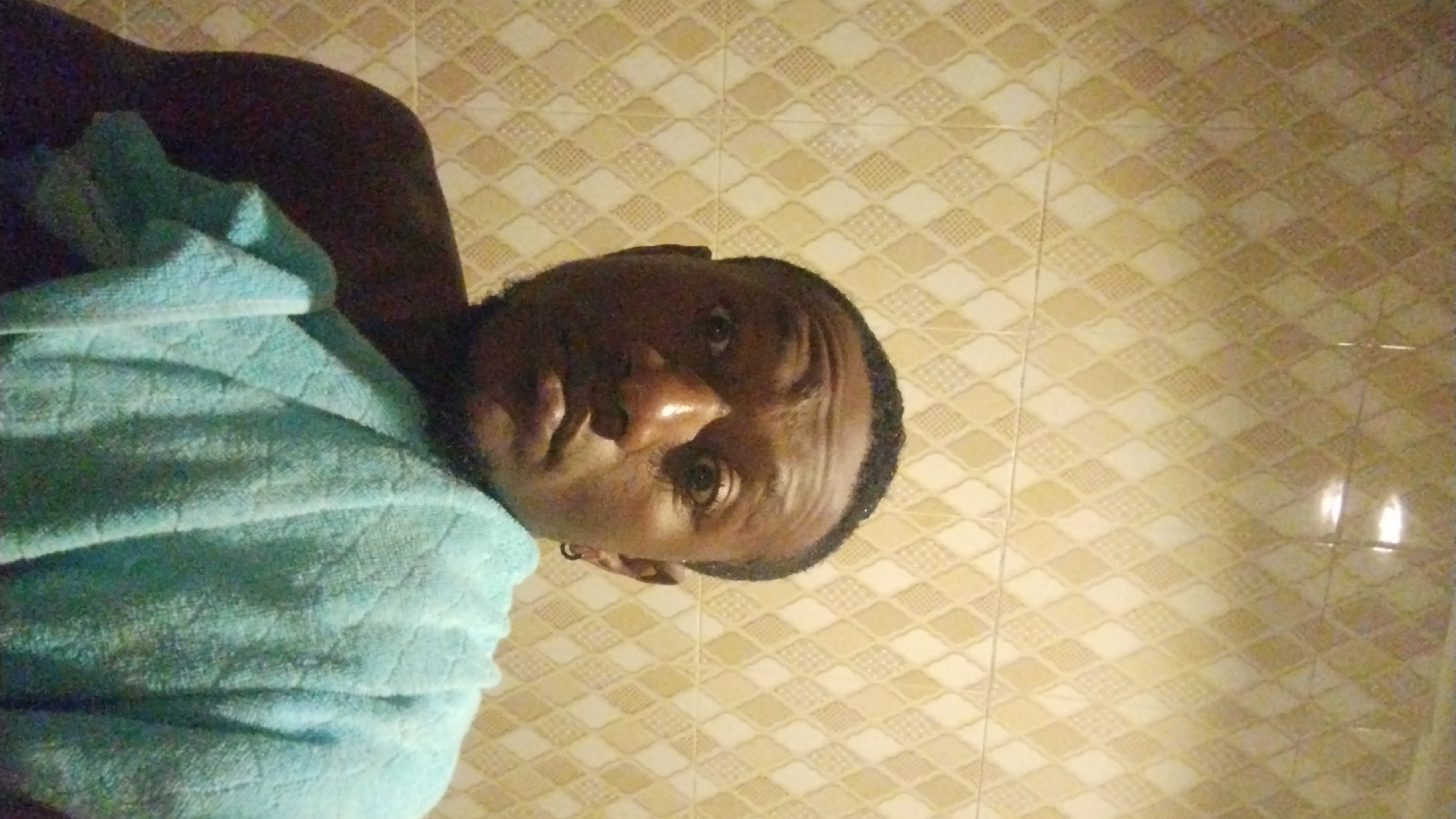 A close-up, dimly lit photograph shows a dark-skinned person with a light blue towel draped over their left shoulder, standing indoors against a beige tiled wall. The person's face is the primary subject, positioned centrally with a slight upward tilt. The wall consists of patterned tiles, featuring a diamond shape design with alternating shades of beige and white. The reflection of light is visible on the wall. There is no visible indication or identifier of Jos, Nigeria. No specific actions or interactions are depicted.