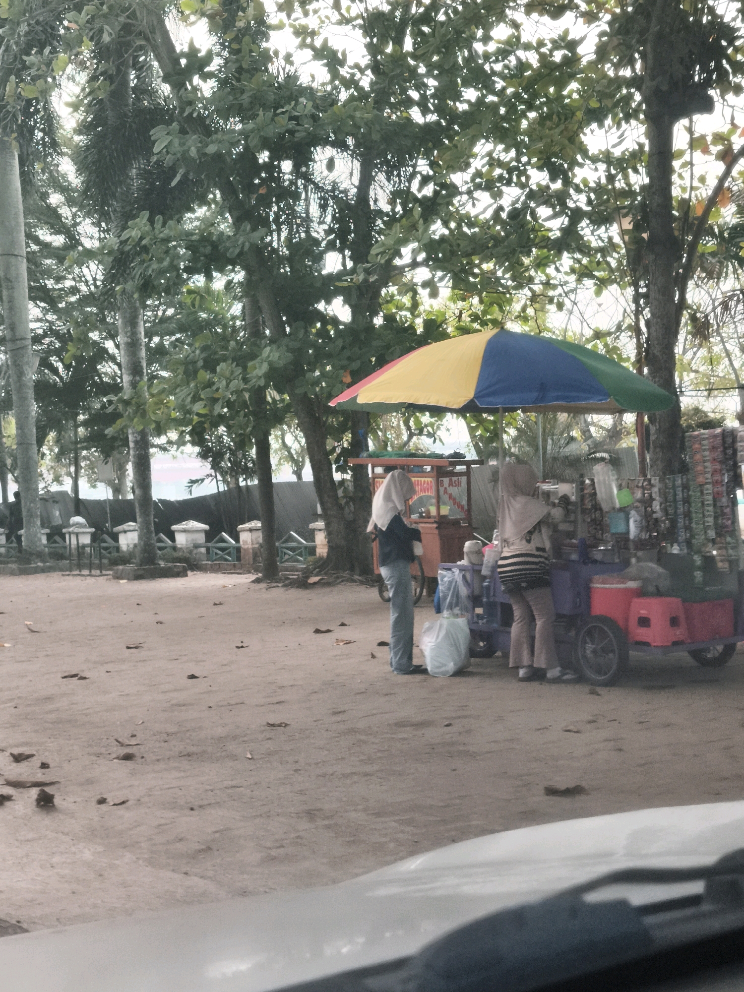 This image depicts a street scene in Palembang, Indonesia, likely near a beach or recreational area given the sandy ground and trees. In the foreground, the hood of a vehicle is visible, suggesting the photo was taken from inside a car.
The main subjects are two individuals standing near food carts. One person, wearing a light-colored hijab and a striped long-sleeved shirt and pants, is interacting with a food cart. Beside them, another person in a white hijab and blue jeans stands near a large white plastic bag.
The food carts are colorful and appear to be selling various items. One cart has a prominent umbrella that is a mix of yellow, blue, and green. Another cart is red and appears to have some signage. A rack of items, possibly snacks or goods, is displayed on the side of one of the carts.
The environment is characterized by sandy ground with scattered leaves. Several trees with lush green foliage are present, providing shade. In the background, a dark barrier or fence can be seen, along with some ornamental pillars, hinting at a structured area beyond the immediate street. The lighting suggests it might be daytime, possibly overcast or early morning/late afternoon.