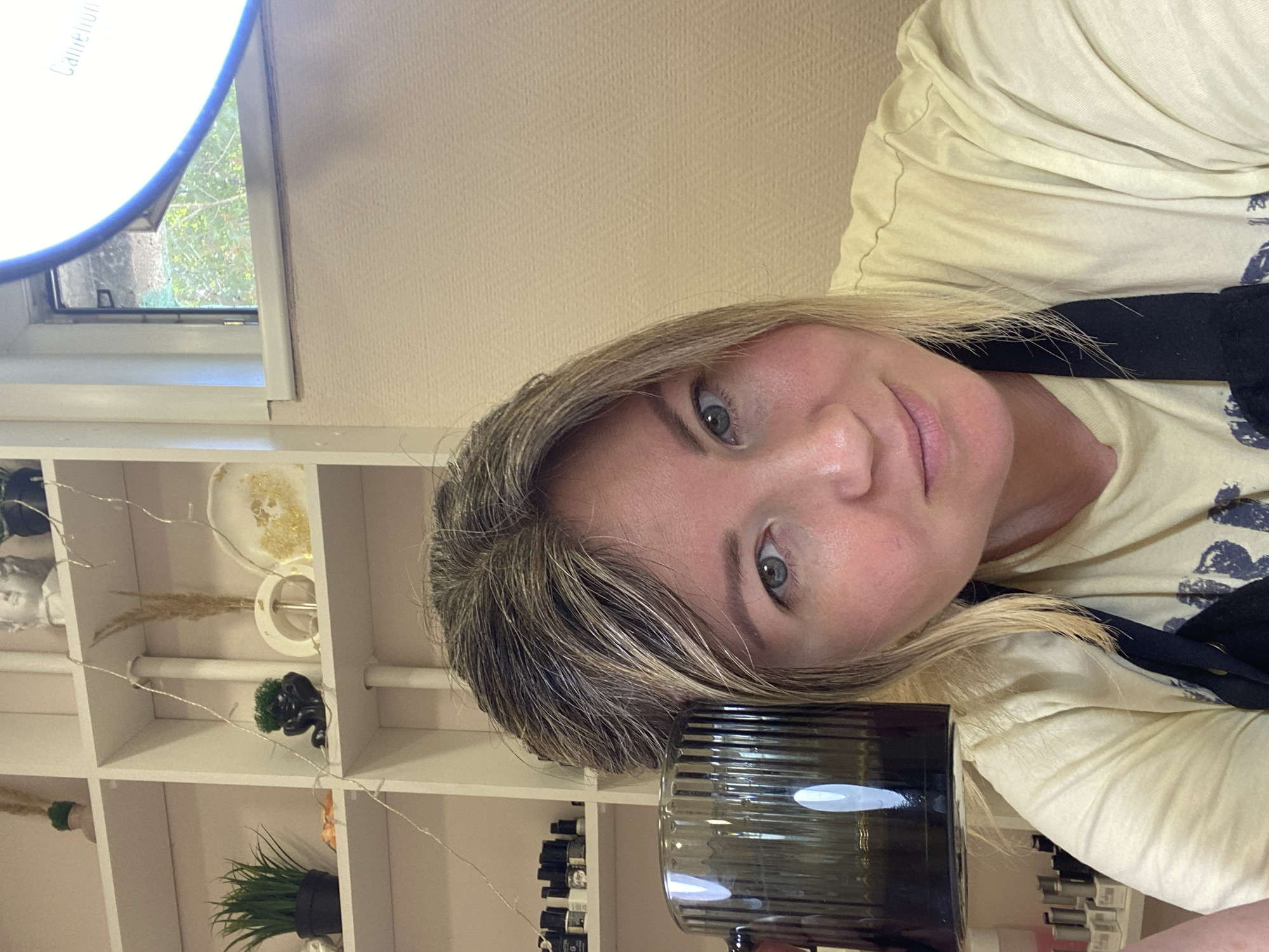 The image shows a woman holding a dark-colored mug to her face, with a shelf of nail polish bottles visible behind her. She is wearing a light-colored t-shirt. The background suggests an indoor setting, possibly a salon or home. The woman is looking towards the camera. The scene does not provide any specific information about the city of Chaikovsky, Russia.