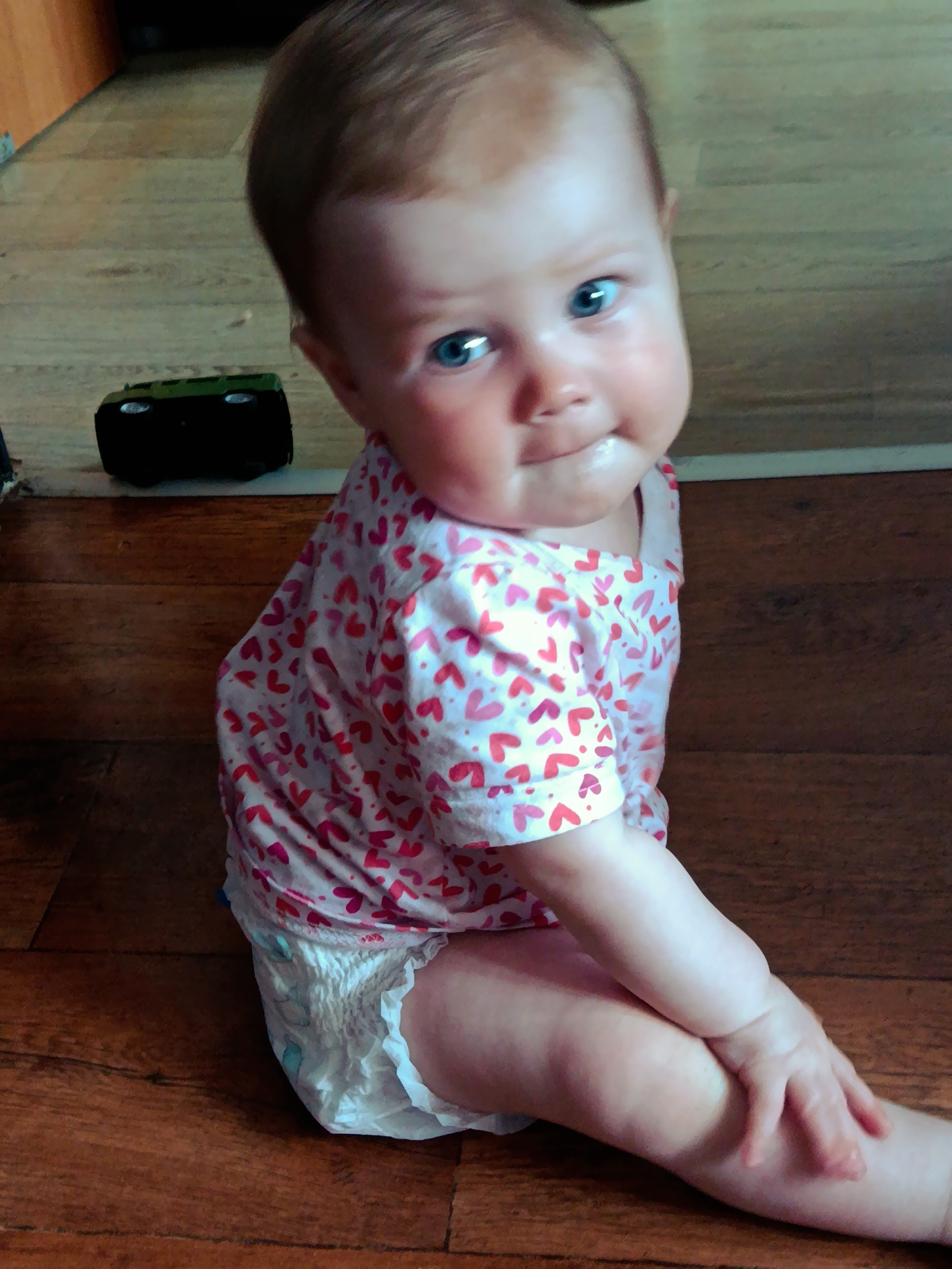 A young baby sits on a wooden floor. The baby is wearing a white shirt with pink hearts and a white diaper. The baby has blue eyes and light brown hair. To the left of the baby is a small black toy car. The location is Фоки, Russia.