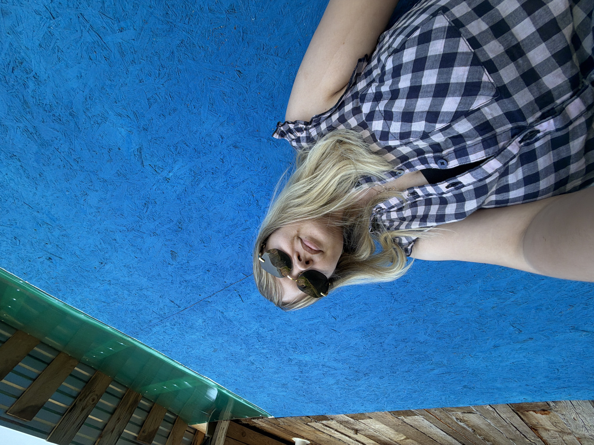 A woman wearing sunglasses and a black and white checkered button-down shirt is lying on a painted blue wood surface. A partially visible structure with green corrugated plastic roofing supported by wooden beams and planks is located in the lower left and lower right corners of the image. The image appears to be taken from a low angle, looking upwards. There is no information given in the image to determine the specific location in the city of Чумна, Russia.