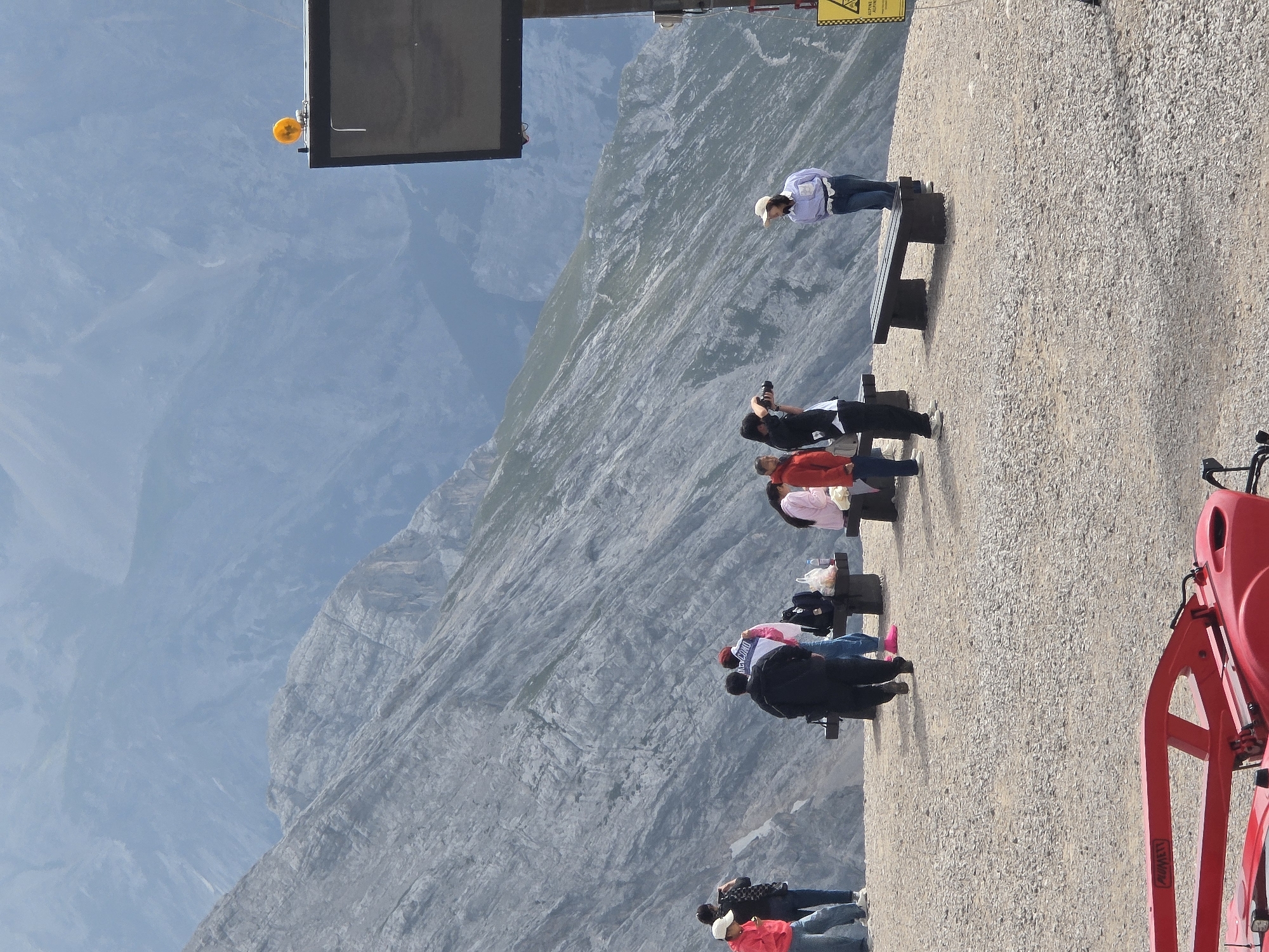 The image depicts a scenic overlook with several people enjoying the mountain view. In the foreground, a red industrial-looking object, possibly a piece of machinery, is partially visible. The ground is covered in gravel. Behind the foreground object, a group of people are spread out on a raised platform. Some are standing, others are seated on benches, and a few are taking photographs.
On the left side of the image, three individuals are visible. One person in a red jacket and white hat is standing, while another in a black jacket is taking a photo. A third person, wearing a red jacket and blue jeans, stands further back.
In the middle ground, several people are gathered around benches. One person in a black jacket and blue jeans is walking away from the camera. Another person in a pink top and white pants is seated. A person in a white and blue jacket is taking a photo, and another person in a pink top is also present.
On the right side of the image, a woman in a light purple shirt and blue jeans is standing, looking towards the camera. A large, dark sign is mounted on a pole behind her, with a small orange light on top. In the far background, a vast, misty mountain range stretches out, with layers of peaks fading into the distance. The overall impression is a popular viewpoint in a mountainous region, likely a tourist destination, possibly in Garmisch-Partenkirchen, Germany, given the mountainous scenery.