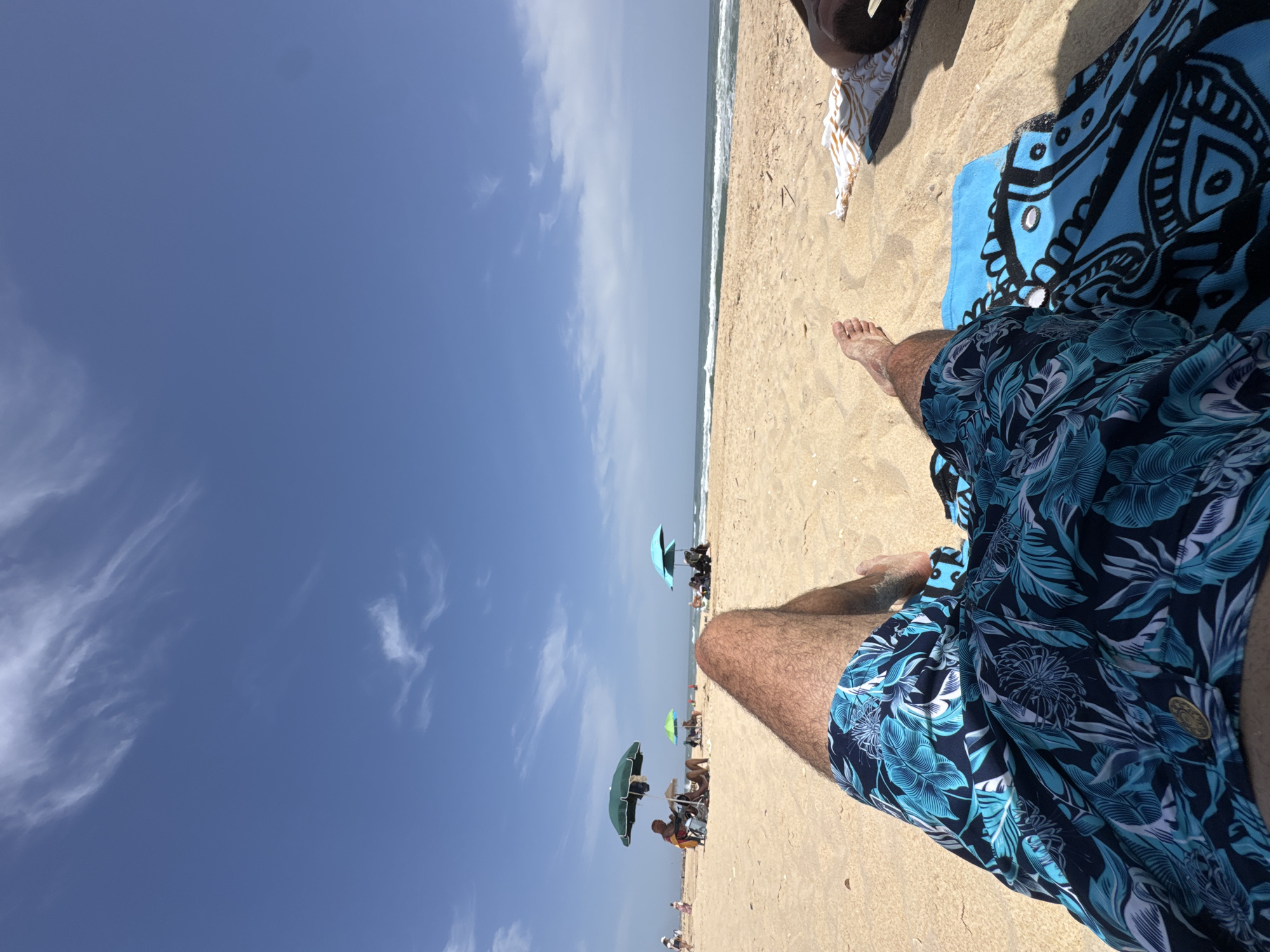 A first-person perspective shot, taken on a beach, shows a pair of legs clad in blue floral swim trunks resting on a sandy surface. A patterned blue and black towel is beside the legs, with a smaller towel nearby. In the distance, other beachgoers sit under umbrellas. The horizon meets the sky, where a few wispy clouds are visible against a clear blue sky. The image is sideways; rotating it counter-clockwise would place the horizon at the top.