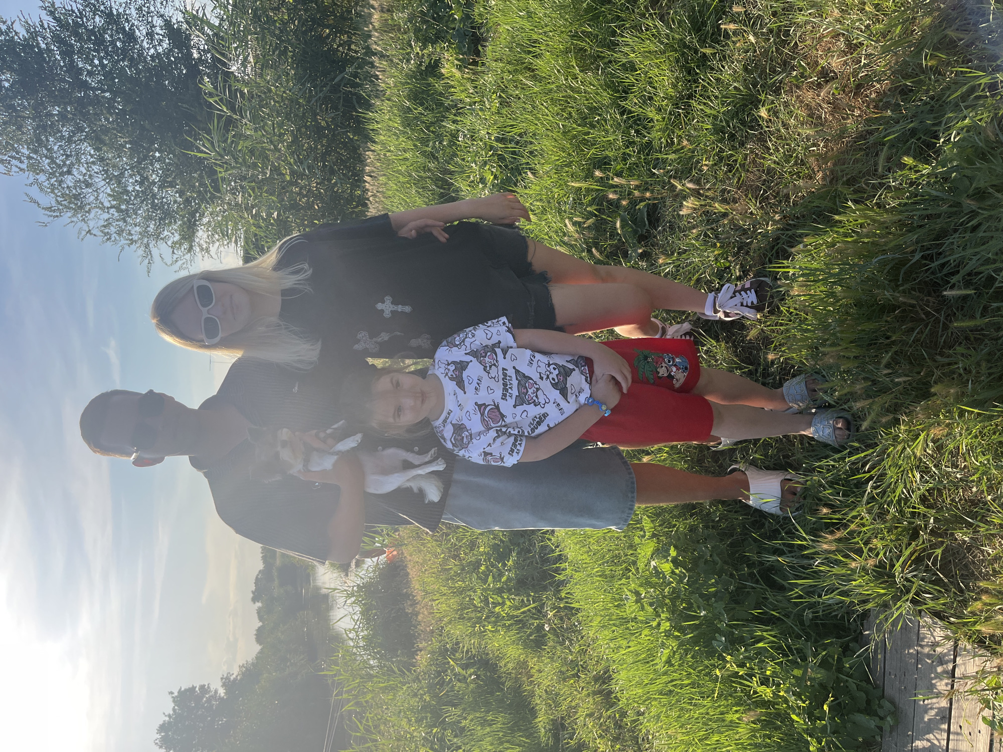 The image captures a family of three and a dog posing for a photograph in a grassy outdoor setting. A man in the center holds a small white and brown dog. He is wearing sunglasses, a dark t-shirt with a graphic print, and light blue denim shorts. To his right stands a young woman with blonde hair, also wearing sunglasses, a dark t-shirt with a white cross graphic, and black denim shorts. In front of the man and slightly to the left is a young girl with dark hair, wearing a white t-shirt with cartoon graphics and red shorts. She has a blue bracelet on her left wrist. All three individuals are looking towards the camera. The background features a body of water on the left, with trees and tall grass. The sky is bright, indicating it is daytime. The overall scene suggests a casual outdoor gathering in a rural or semi-rural environment, possibly near a body of water. The location is identified as Yanovo-Grushevsky, Russia.