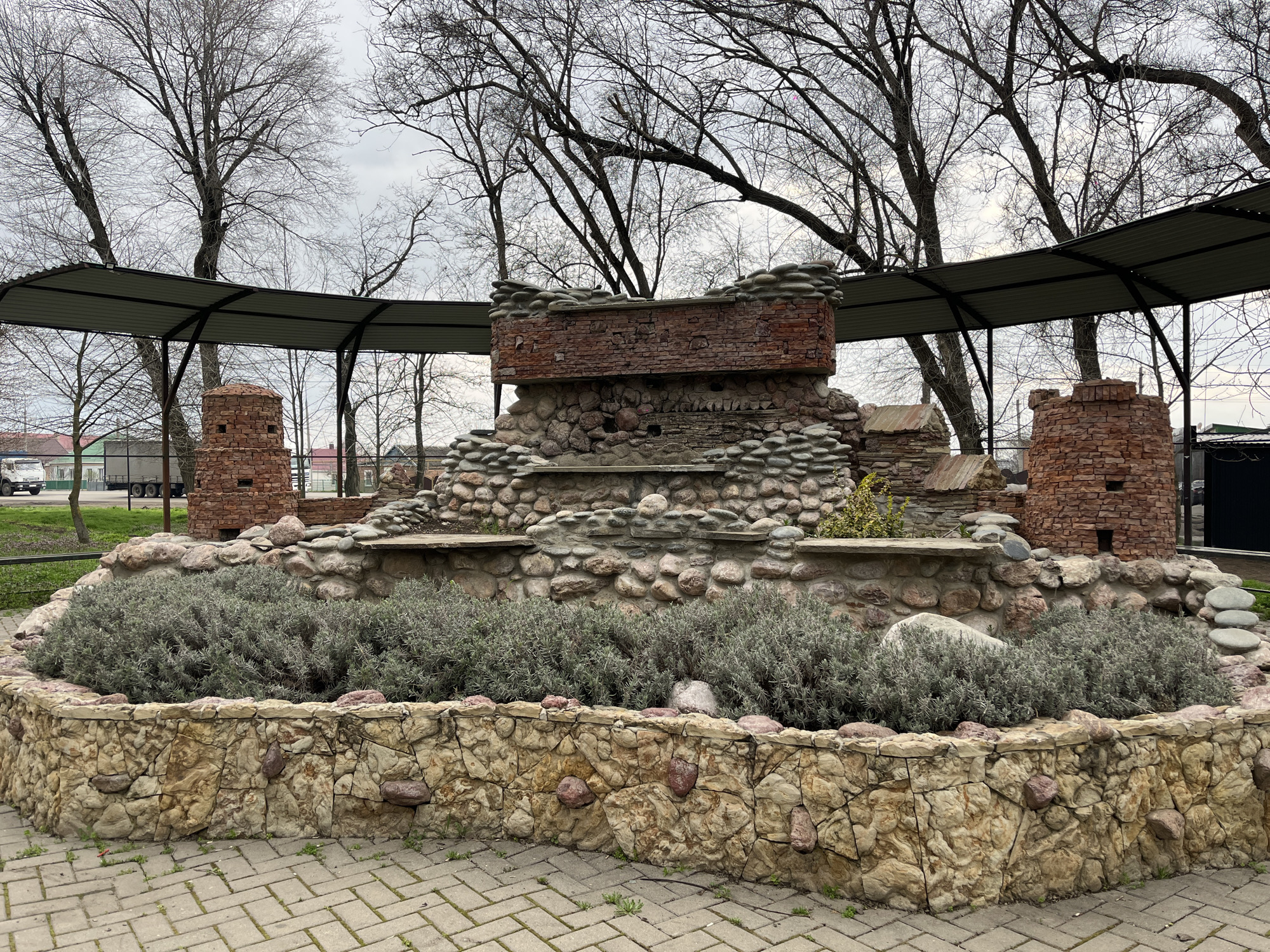 The image shows a decorative structure in Novopokrovskaya, Russia. It features a stone and brick fountain or planter with tiered levels, topped with a brick structure resembling a ruined building fragment. The structure is enclosed by a low wall constructed from fitted stones, and the central area is filled with grey-green plants. Two decorative brick towers flank the central fountain piece. The entire display is covered by a curved metal roof or canopy supported by posts, suggesting a park or public space. The background includes bare trees, a building, and a glimpse of a truck. The ground is paved with interlocking paving stones. The sky appears overcast.