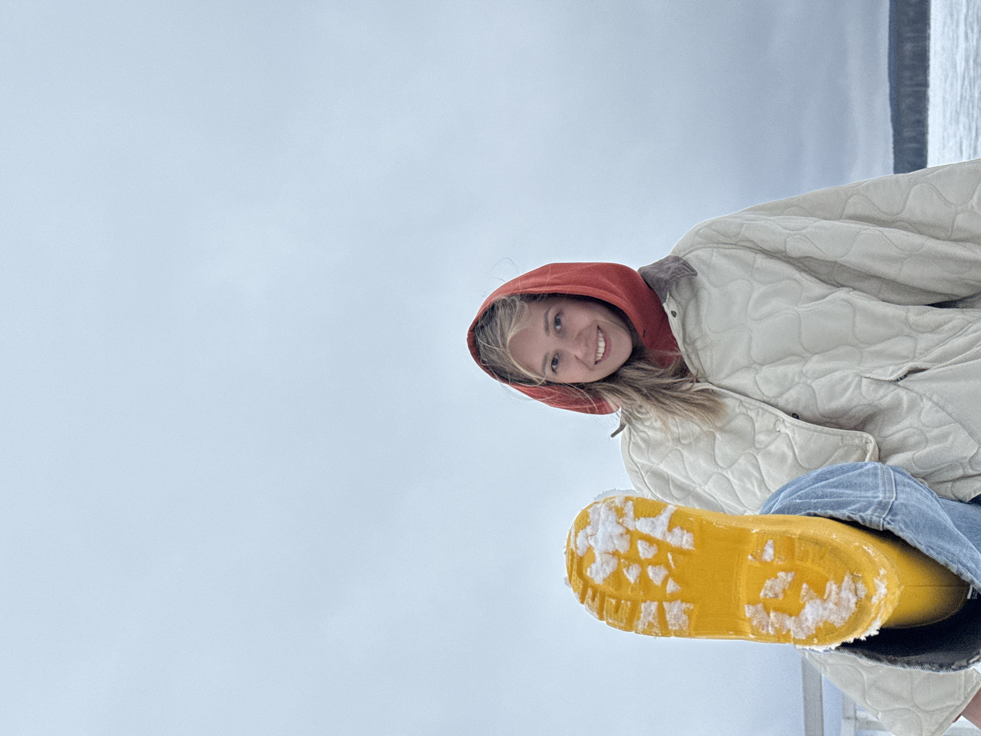 A young woman is pictured outdoors, likely in Chaikovsky, Russia, given the prompt's context. The image appears to be shot from a low angle, looking up at the subject. She is smiling and looking at the camera, wearing a red hooded sweatshirt under a light-colored, quilted jacket and blue jeans. She is holding out a yellow rubber boot, the sole of which is visible and covered in snow. The background consists of a cloudy sky and what appears to be a body of water in the distance. The overall impression is of a casual, informal snapshot.