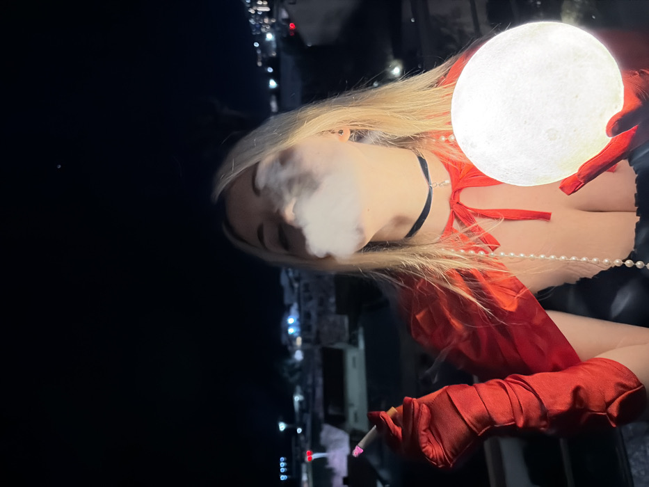 A blonde woman exhales smoke, dressed in red gloves and a matching red top with a black choker and a string of pearls. She holds a lit cigarette in one hand and a glowing orb resembling the moon in the other. The background features a night scene with a cityscape.