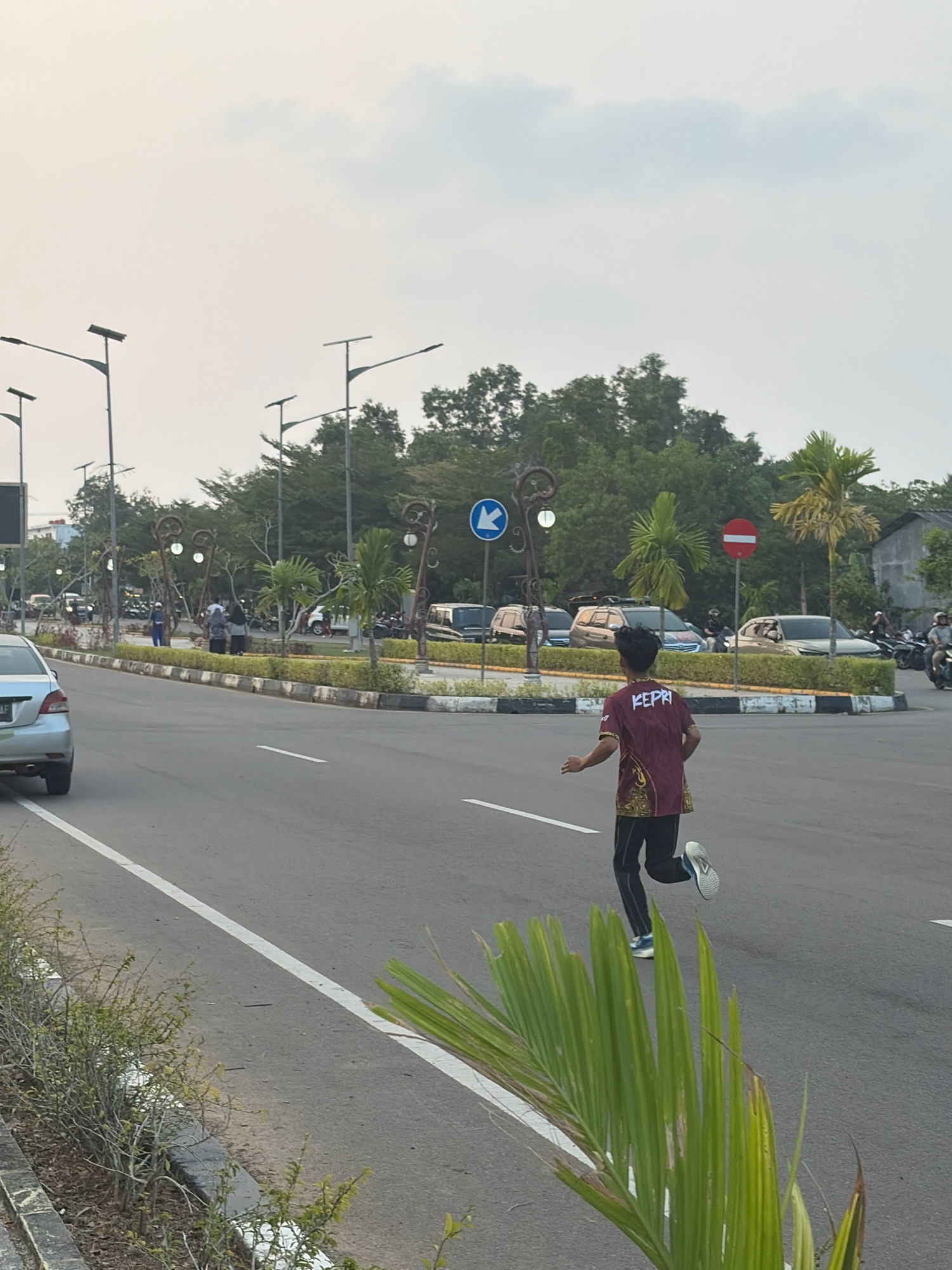 A young male wearing a maroon shirt with gold and black patterns and the word "KEPRI" in white lettering, black pants, and white and blue athletic shoes is captured mid-stride running across an asphalt road in Tanjung Pinang, Indonesia. The road features a white lane divider and a curb with a decorative border. In the background, several parked cars and motorcycles are visible on the opposite side of the road, along with a median strip featuring palm trees and decorative street lamps. A blue circular sign with a white arrow pointing left and a red circular sign with a white horizontal bar are also present. The sky is overcast, suggesting either early morning or late afternoon. The foreground shows a section of a green plant with long, thin leaves.