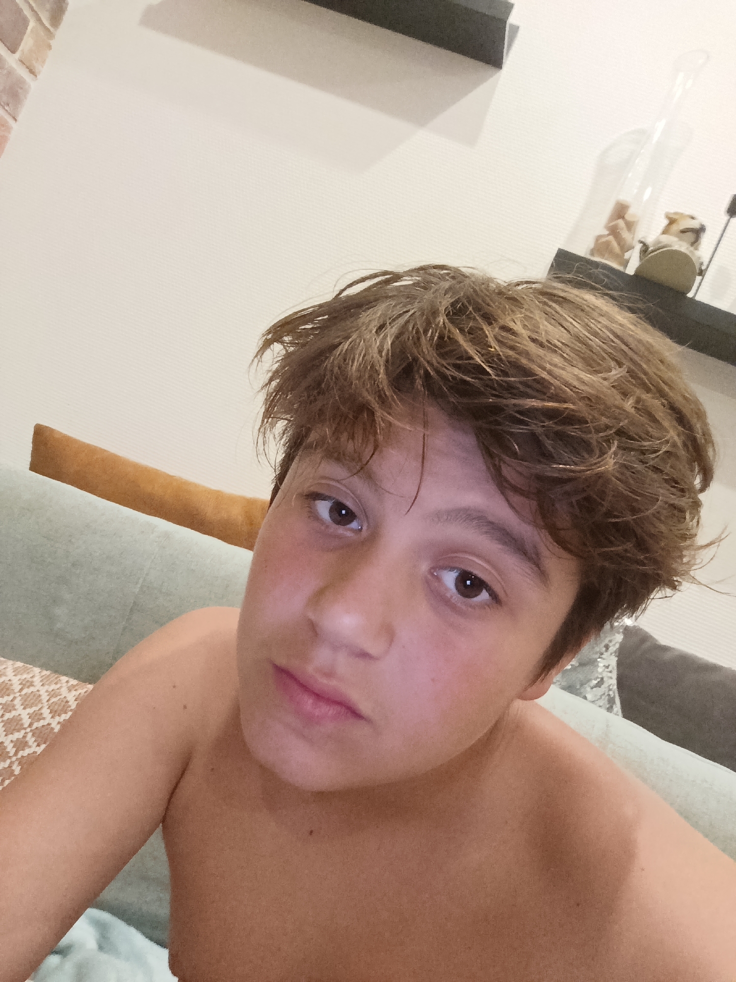 A close-up, chest-up selfie shows a young person with tousled brown hair. They have brown eyes and fair skin with some redness on their cheeks. The person is shirtless and appears to be relaxed, possibly at home, given the background elements. Behind them, a light-colored wall is visible, with a dark shelf mounted on it. On the shelf, a glass jar with some light-colored objects inside and a small figurine can be seen. A light green sofa with a brown decorative pillow is also partially visible. The lighting suggests it might be evening or indoors. The setting appears to be a residential space in Засечное, Russia. No visible text is present in the image.