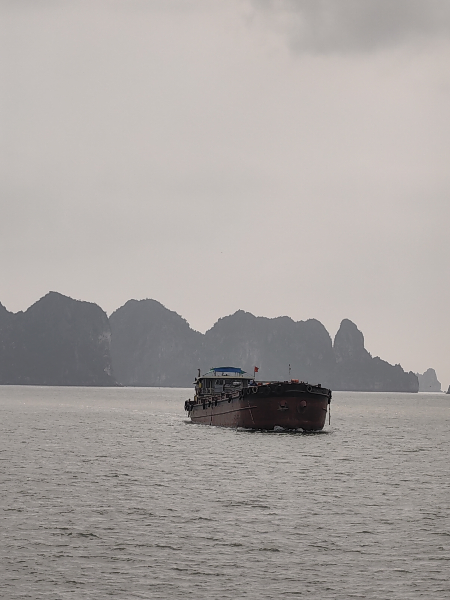 A medium-sized, rust-colored cargo ship is centered in the frame, floating on the gray water of Ha Long Bay, Vietnam. The ship has a small cabin painted blue and white. A small Vietnamese flag is seen on the cabin. In the background, dark-gray, limestone cliffs with sparse vegetation rise out of the water. The overcast sky is a lighter shade of gray. The water in the foreground displays a slight ripple effect. The image was taken in Tuan Chau Ward, Vietnam.