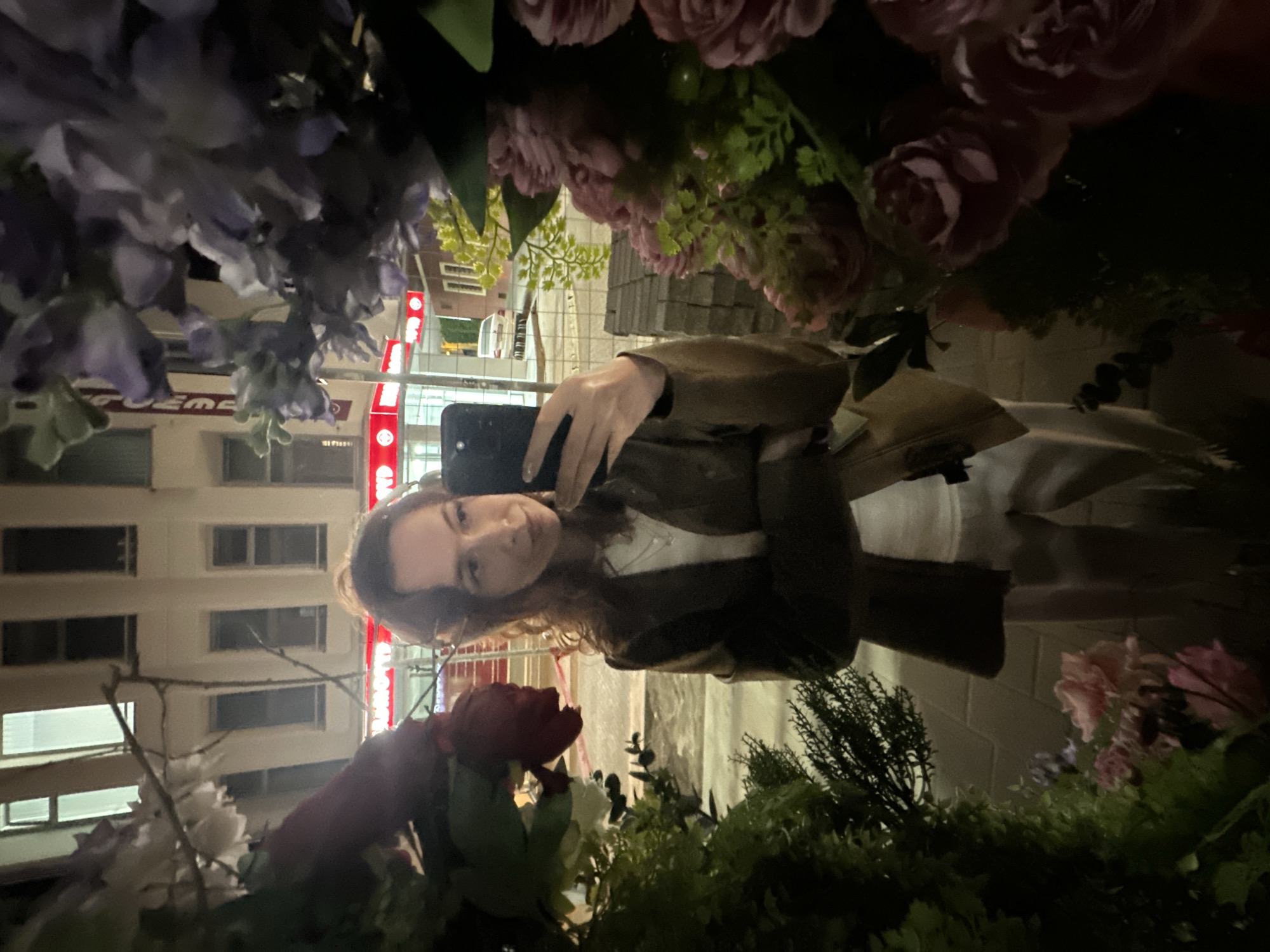 A woman with curly light brown hair is holding a smartphone in her left hand and taking a selfie among many artificial flowers in Novi Sad, Serbia, at night. The woman is wearing a brown leather jacket and a white top. The background includes a building with rows of windows and a red sign with white lettering. The lighting suggests it's either very late or early morning. The flowers include shades of purple, pink, red, white, and green.