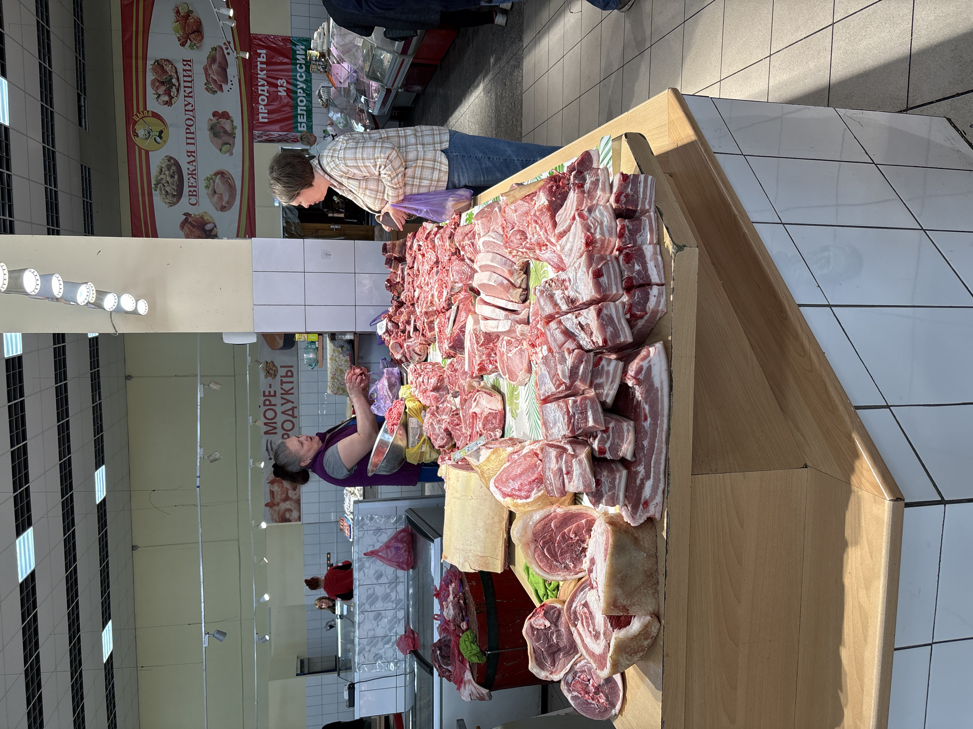 This image depicts a butcher shop, likely in Mezhdurechensk, Russia, given the Cyrillic script on signage. The foreground is dominated by a wooden display case piled high with various cuts of raw meat, including what appears to be pork ribs, bacon, and other unidentified cuts. A female butcher, wearing a purple top, stands behind the counter, seemingly attending to customers. One customer, a woman wearing a plaid shirt, is visible leaning over the counter, possibly making a purchase. The butcher's workspace behind the display case includes a metal scale and a tiled wall. In the background, other vendors and customers can be seen in the market setting. The lighting is provided by overhead fluorescent fixtures. Signage with images of meat products and the words "Свежая продукция" (Fresh Products) and "из Белоруссии" (from Belarus) is visible above the counter.