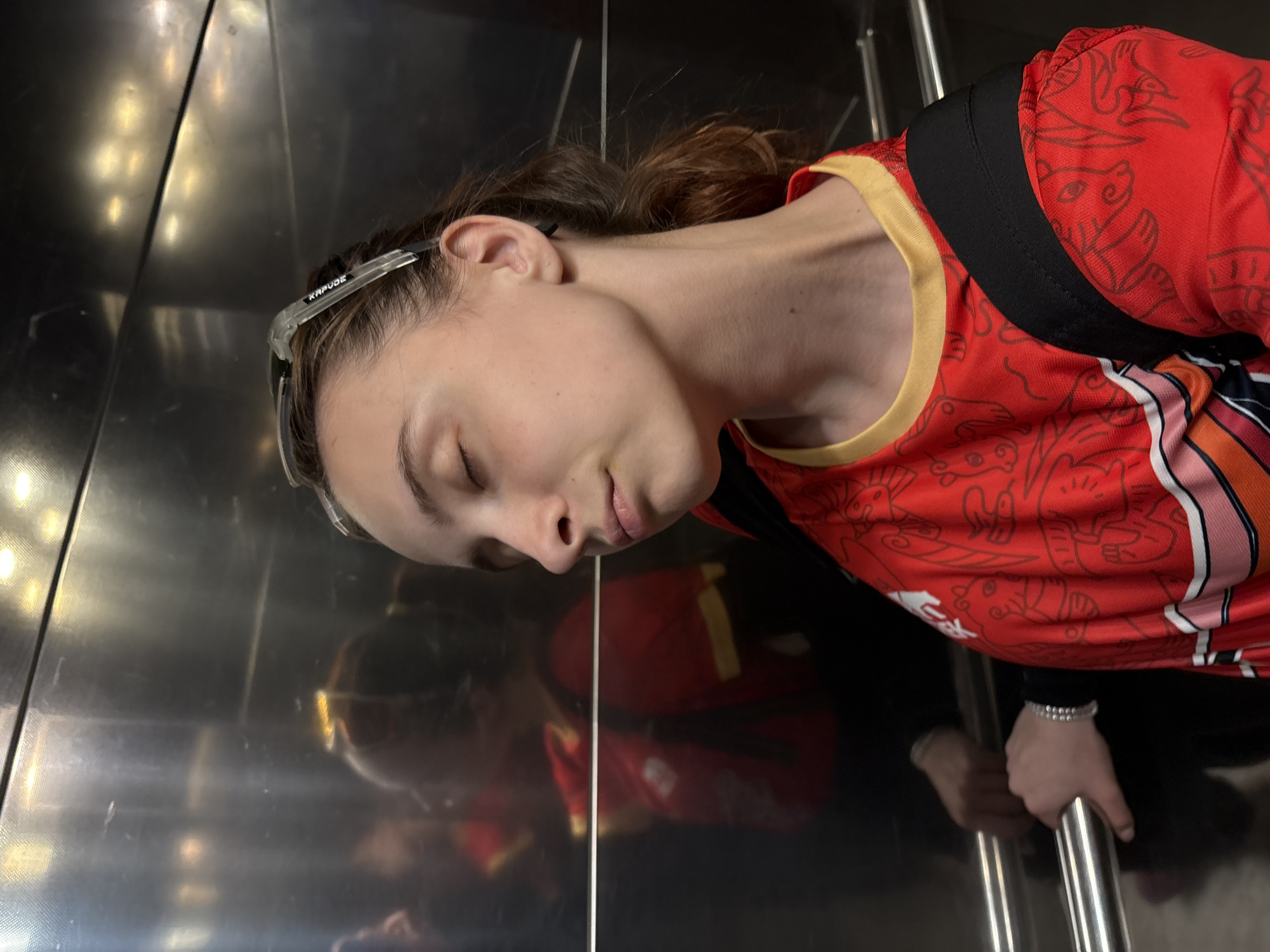 A woman with light skin, closed eyes, and brown hair is positioned in an elevator. She is wearing a red t-shirt with orange and white accents, a black backpack strap over her shoulder, and clear-framed glasses resting on her head. Her right hand is holding onto a metal bar. The elevator's interior has a metal wall reflecting the woman and her clothing. The image was taken in the city of Perm, Russia.