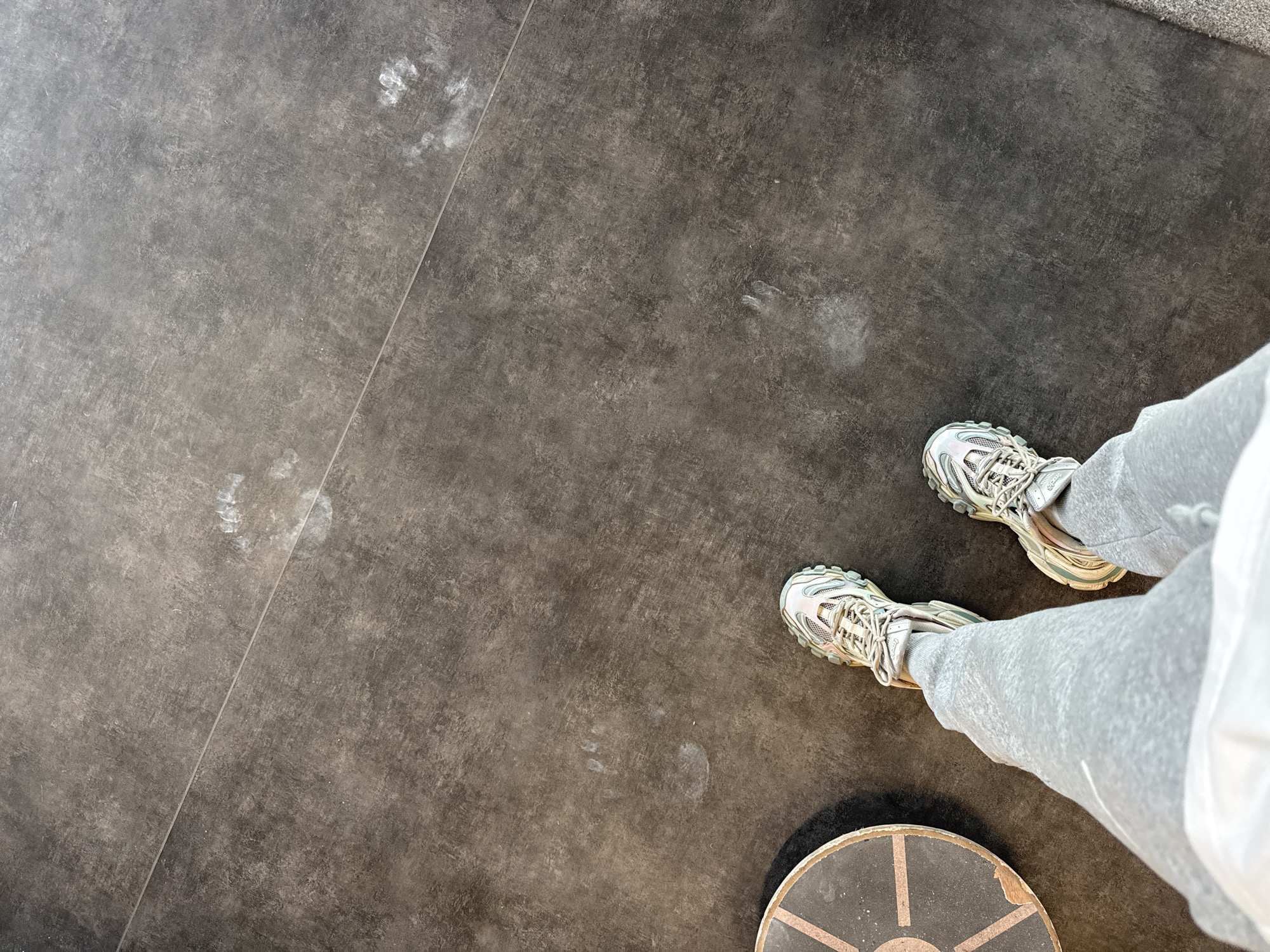 The image shows a person standing on a gray, tiled floor. The person is wearing gray sweatpants and iridescent sneakers. A wooden balance board is partially visible in the lower right corner of the image. There are several white, smeared footprints on the floor in the upper left quadrant. The location is most likely indoors. There is no information in the image to confirm the location is Sofia, Bulgaria.