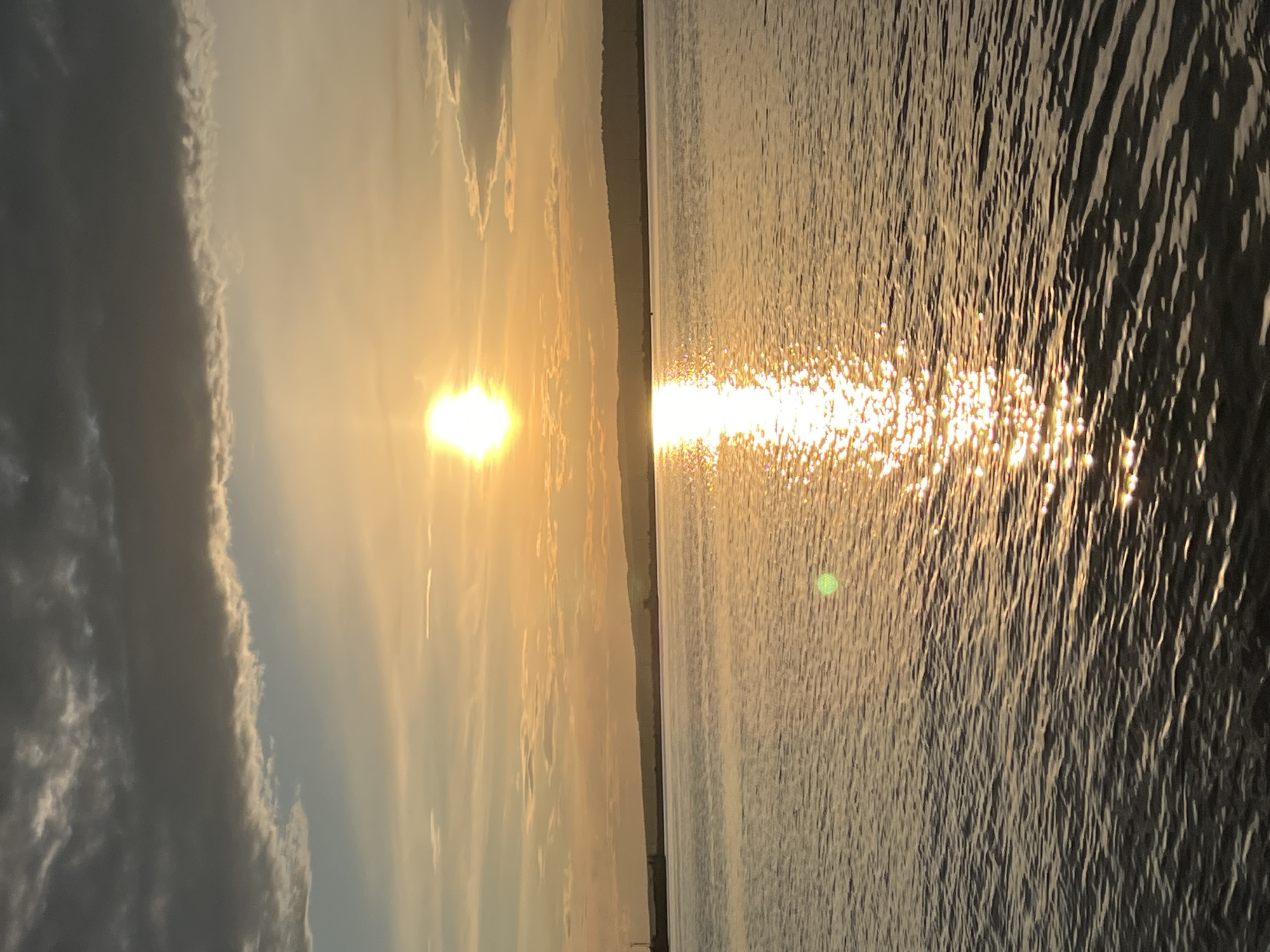A serene sunset scene unfolds over the Kama River in Chaikovsky, Russia. The sun, a brilliant orb of light, hovers near the horizon, casting a golden glow across the water. Its reflection shimmers on the rippling surface, creating a dazzling pathway of light. Dark clouds drift across the sky, tinged with orange and yellow hues from the setting sun. In the distance, a dark, silhouetted treeline marks the far bank of the river. The water is calm, with gentle waves reflecting the light and sky. The overall atmosphere is one of tranquility and natural beauty.
