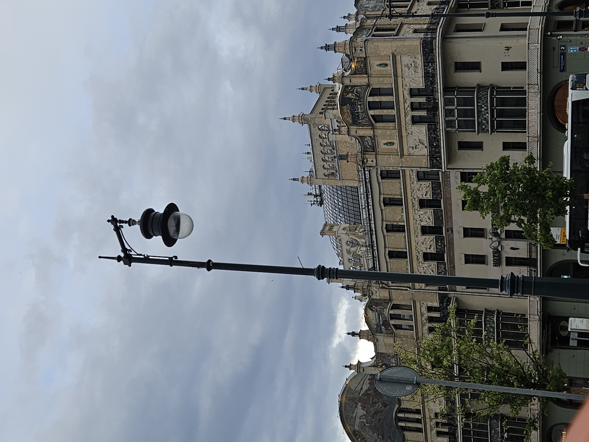 The image shows a street scene in Moscow, Russia, with a focus on architectural details. A black lamppost stands prominently in the foreground against a cloudy sky. Behind the lamppost is an ornate, multi-story building with detailed facade work and spired roofs. A tree partially obscures the building’s mid-section, and a bus is visible on the right side of the frame. A street sign is seen in front of the building to the lower-left.