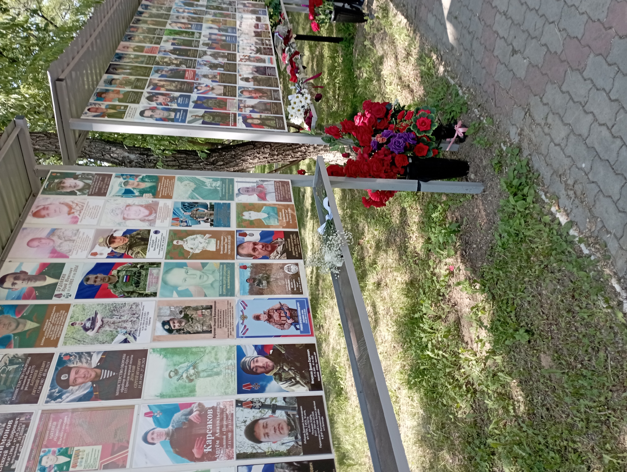 The image shows a memorial display of portraits, likely of fallen soldiers, arranged on metal stands outdoors. The portraits are laminated and displayed in rows. The memorial is situated in an outdoor setting with grass, trees, and a paved walkway visible. Floral tributes, including red and purple flowers, are placed at the base of the display. The overall atmosphere is solemn and commemorative. The text on some of the portraits is in Russian. Based on visual cues and common memorial practices in Blagoveshchensk, Russia, this memorial is likely located at the Alley of Heroes in the city.